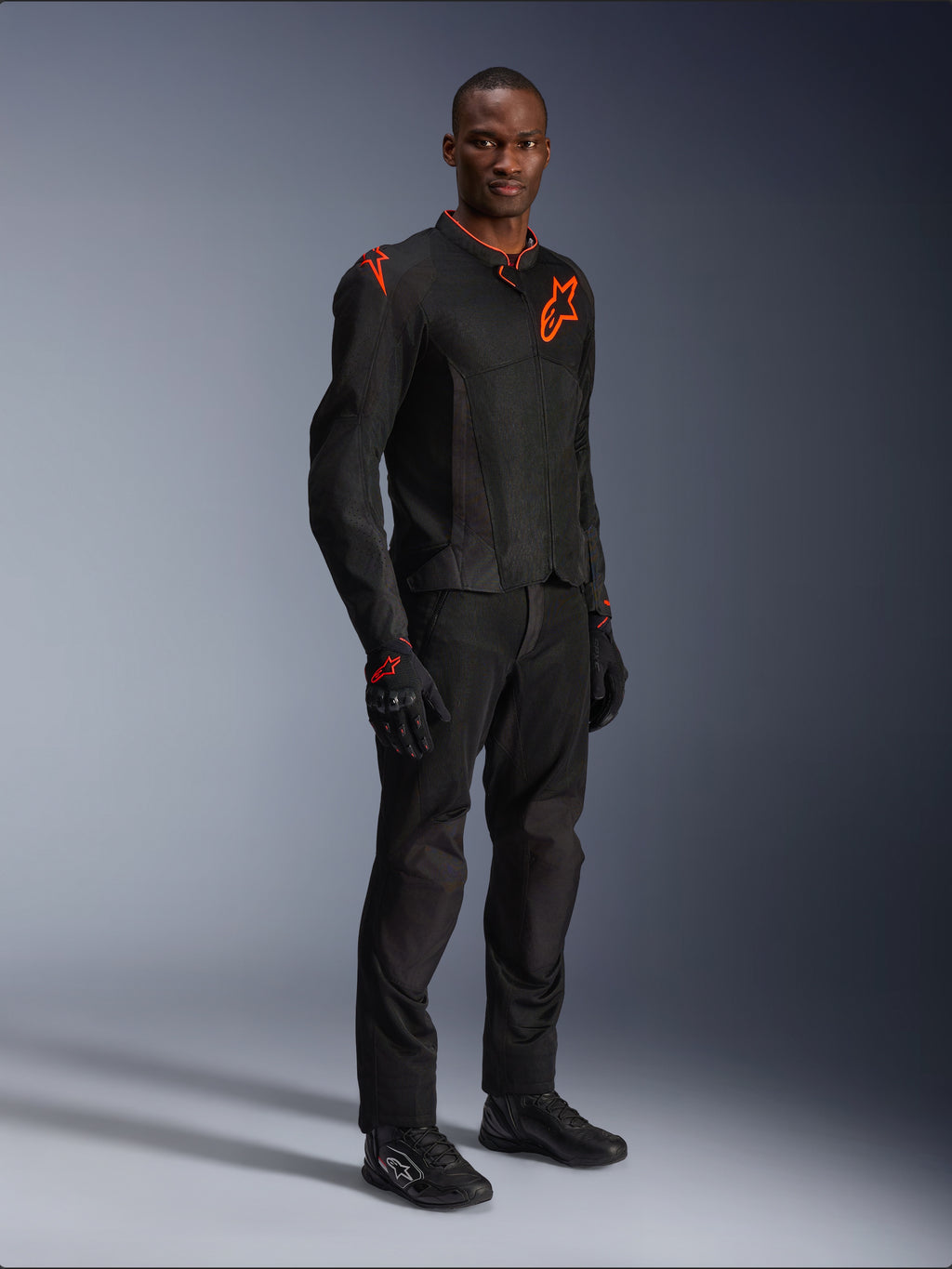 Modèle portant la veste Alpinestars Viper Air V4 en noir et rouge fluo, dotée de panneaux en mesh et de logos étoilés rouges sur la poitrine et l'épaule, accompagnée d'un pantalon de moto, de gants et de chaussures noirs assortis, ensemble d'équipement de route professionnel