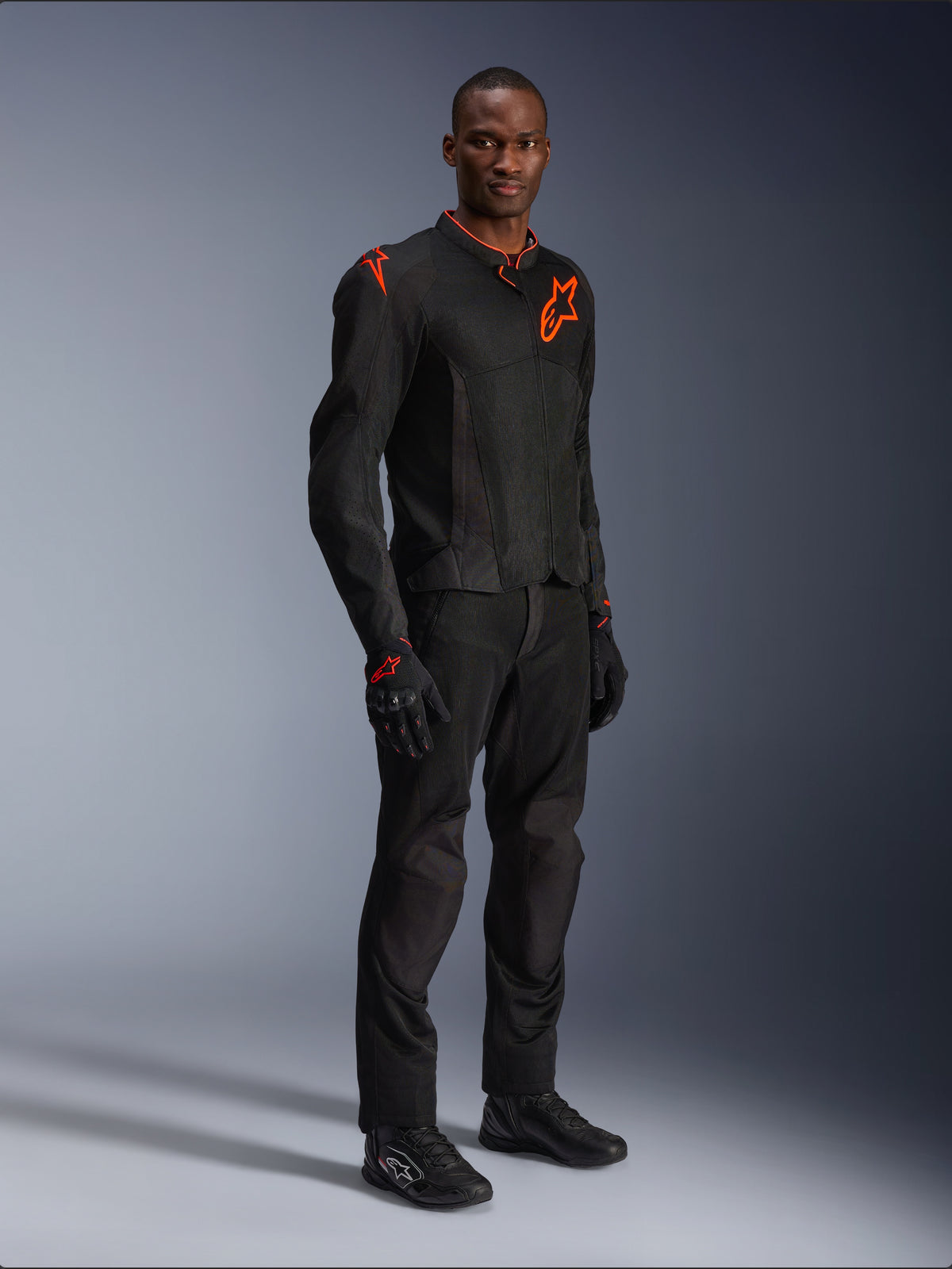 Model in der Alpinestars Viper Air V4 Jacke in Schwarz und Red Fluo, mit Mesh-Einsätzen und roten Stern-Logos auf der Brust und Schulter, kombiniert mit passender schwarzer Motorradhose, Handschuhen und Fahrschuhen, professionelles Motorrad-Outfit für die Straße