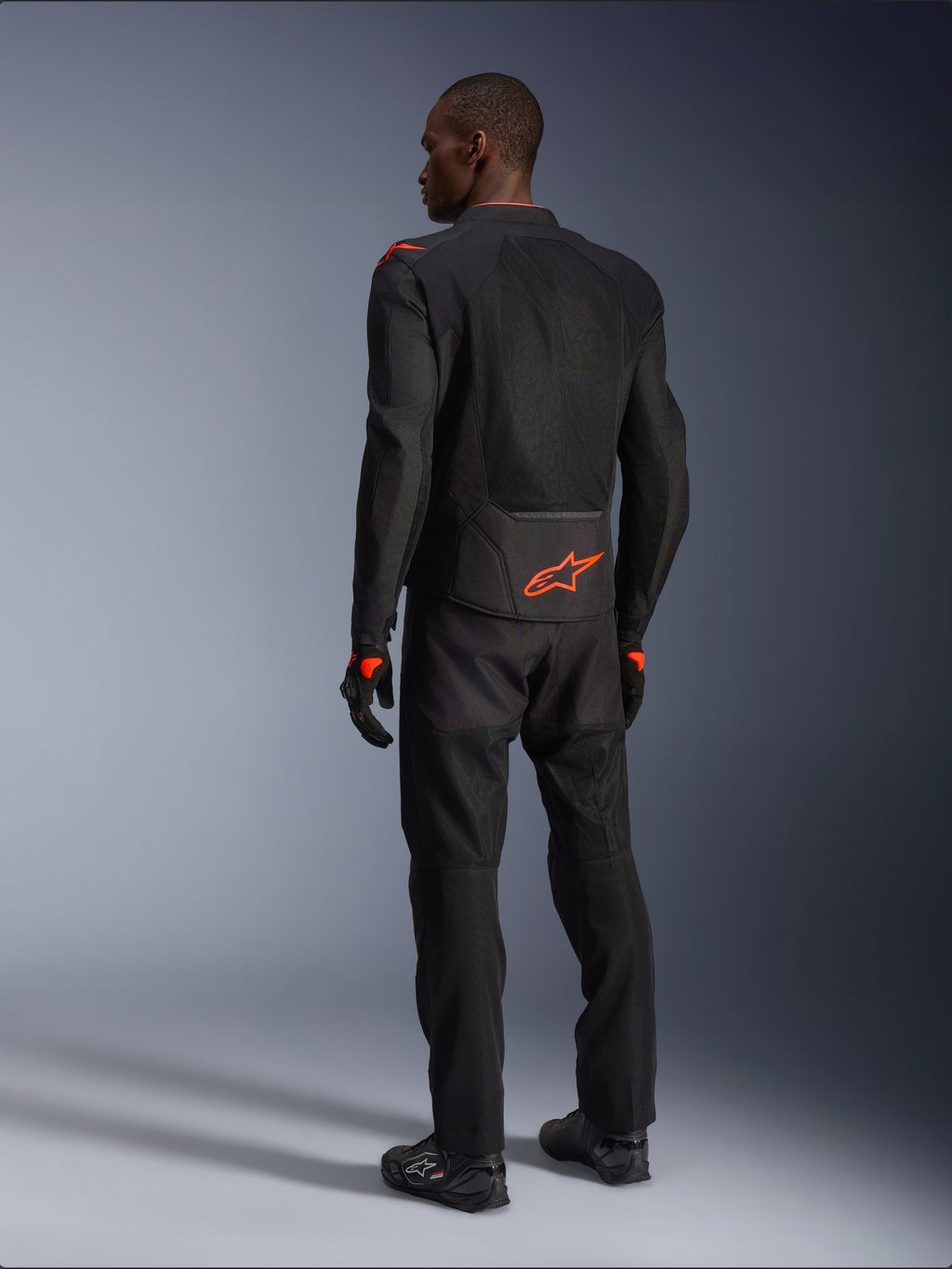 Pilote homme vu de dos portant la veste Alpinestars Viper Air V4, noir et rouge fluo, logo Astars orange sur le bas du dos et les épaules, construction en textile mesh, avec pantalon et bottes de moto noirs assortis