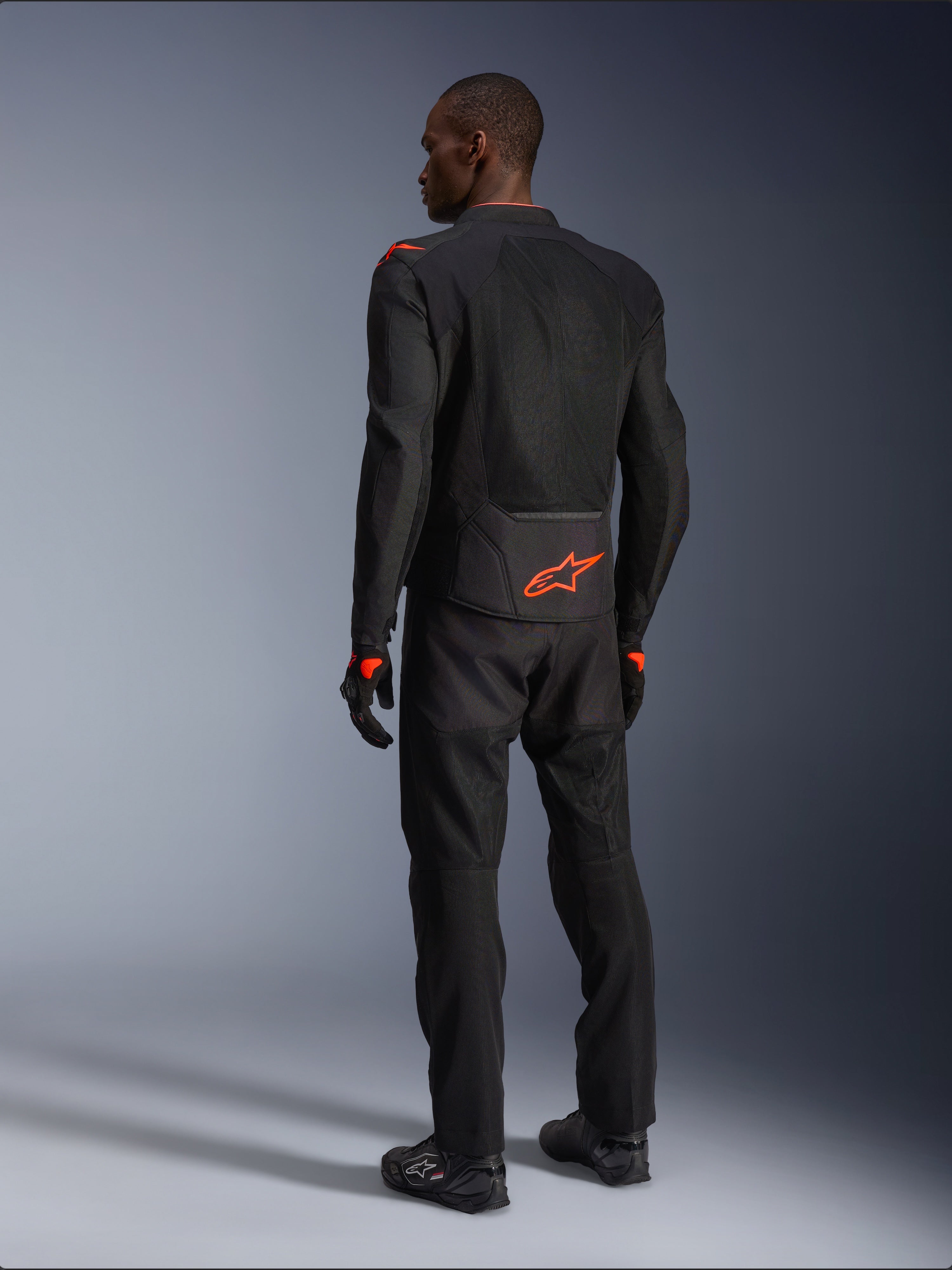 Pilote homme vu de dos portant la veste Alpinestars Viper Air V4, noir et rouge fluo, logo Astars orange sur le bas du dos et les épaules, construction en textile mesh, avec pantalon et bottes de moto noirs assortis