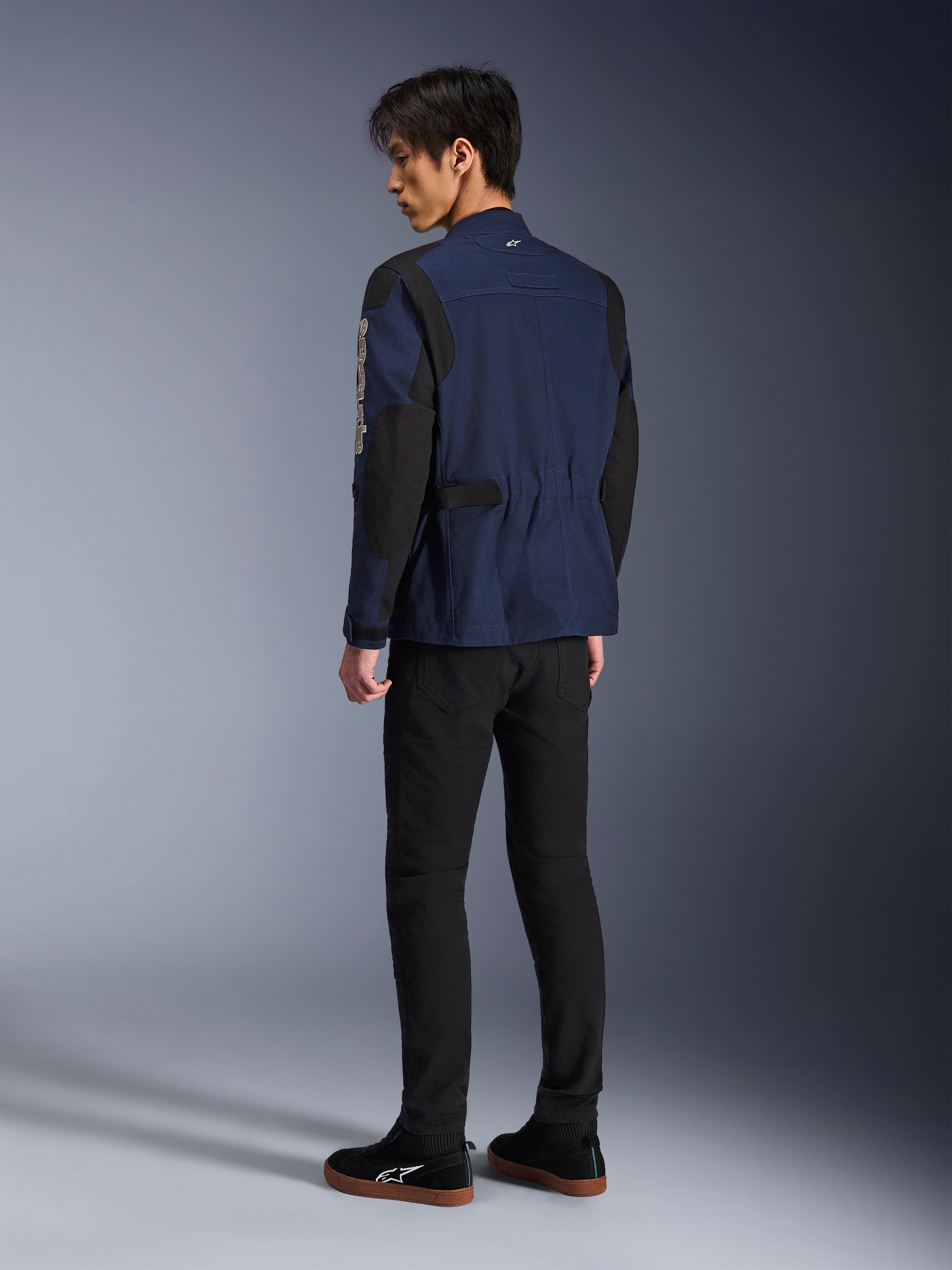 Männliches Model in der Rückansicht trägt die Alpinestars Dice Jacke in Dunkelblau und Schwarz, kombiniert mit einer schwarzen Hose und schwarzen Sneakern, urbane Motorrad-Fieldjacket mit Schulterfalten und verstellbaren Taillenriegeln.