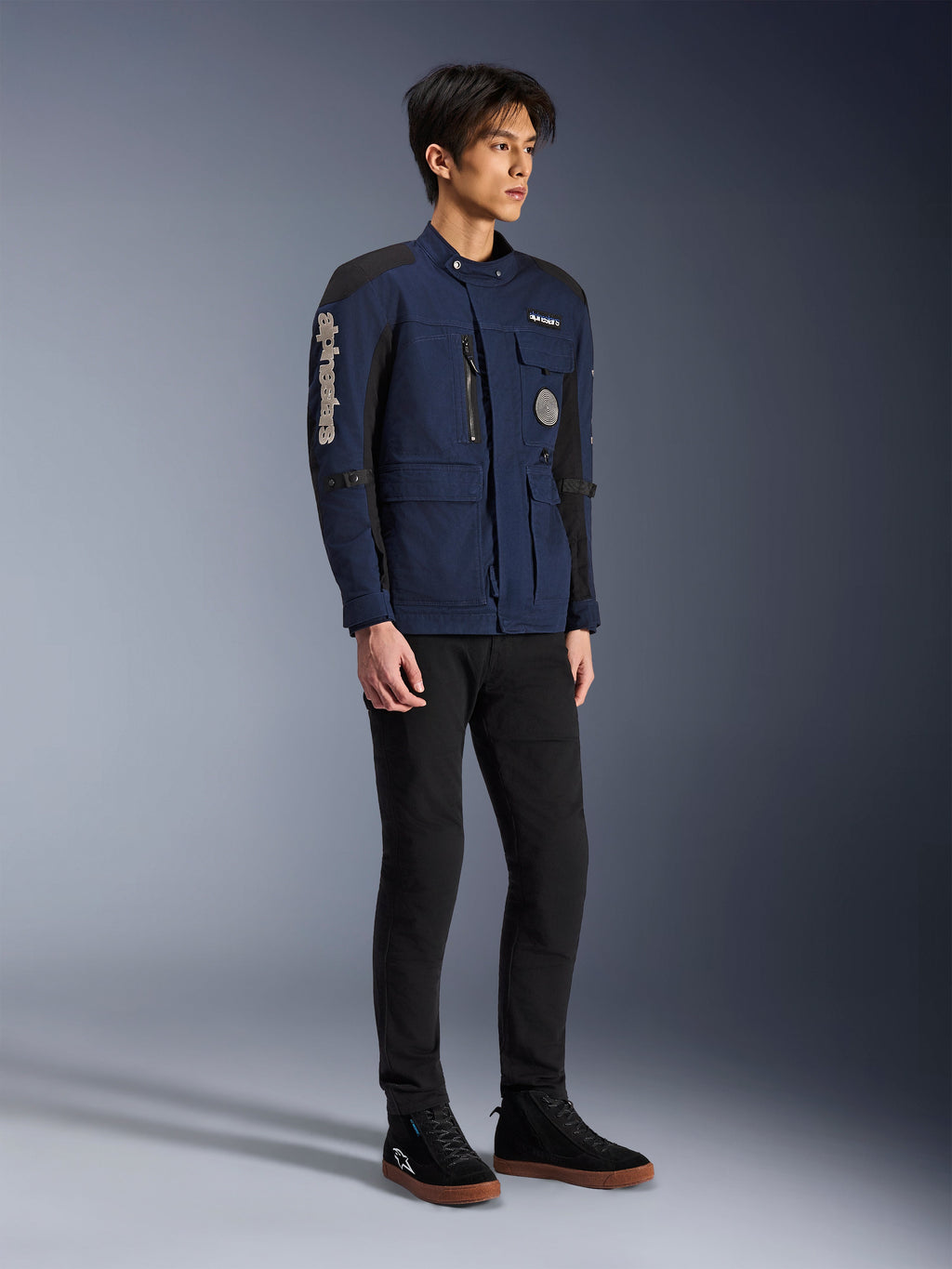 Alpinestars Dice Jacke, Dunkelblau und Schwarz, moderne Motorradjacke im Field-Style getragen von einem Model mit schwarzer Hose und schwarzen High-Top-Sneakern, mit mehreren Utility-Taschen, vertikalem Alpinestars-Branding am Ärmel und gewebten Aufnähern, urbane zeitgemäße Fahrerausrüstung