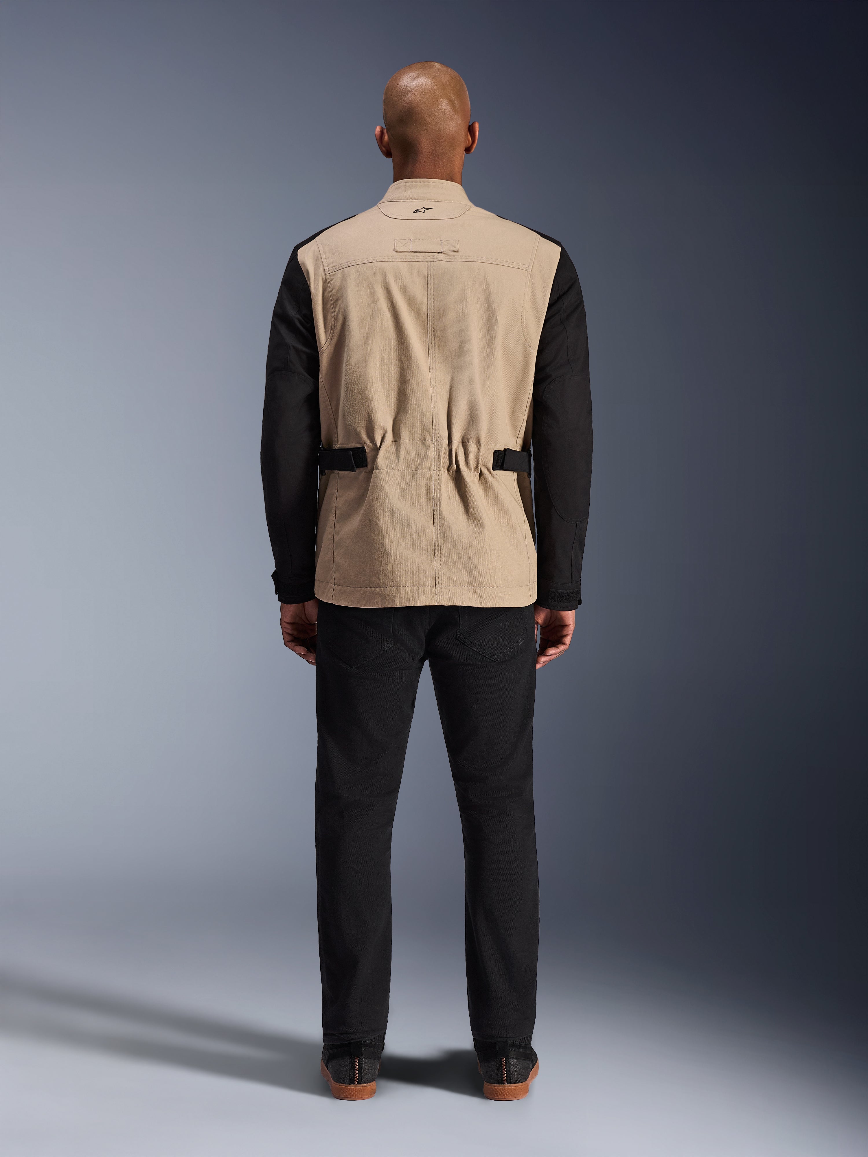 Veste Dice Alpinestars, veste de moto urbaine, beige et noir, vue de dos sur mannequin, dotée d'une construction en sergé de polycoton, de sangles de réglage à la taille et d'un petit logo brodé sur le col, portée avec un pantalon noir