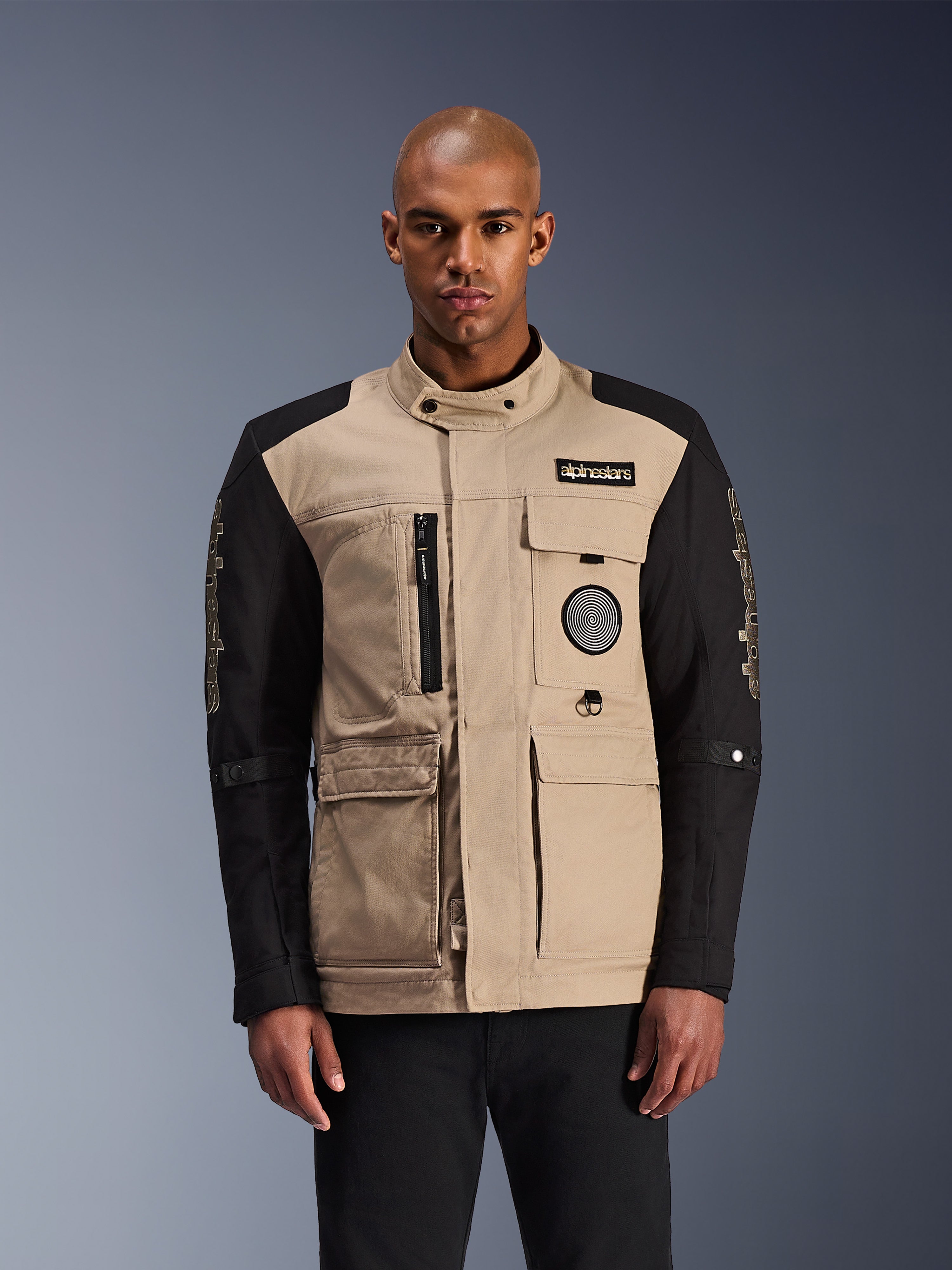 Alpinestars Dice Jacke, urbane Motorradjacke, Beige und Schwarz, robuste Workwear-Ästhetik mit mehreren Funktionstaschen und Brust-Patches, hoher Kragen mit Knopfverschluss, markantes Branding an den Ärmeln, getragen von einem männlichen Model