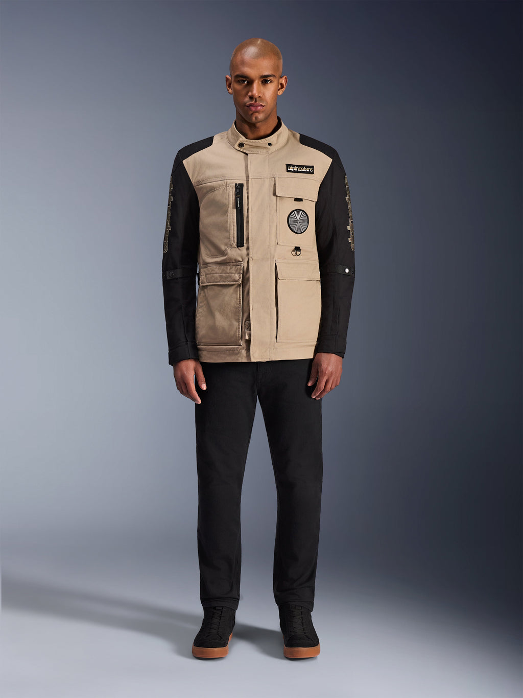 Veste Alpinestars Dice, veste de moto urbaine, corps beige aluminium avec manches noires, silhouette inspirée du workwear avec quatre poches utilitaires frontales, portée avec un pantalon noir et des baskets montantes noires, équipement de moto robuste de style urbain