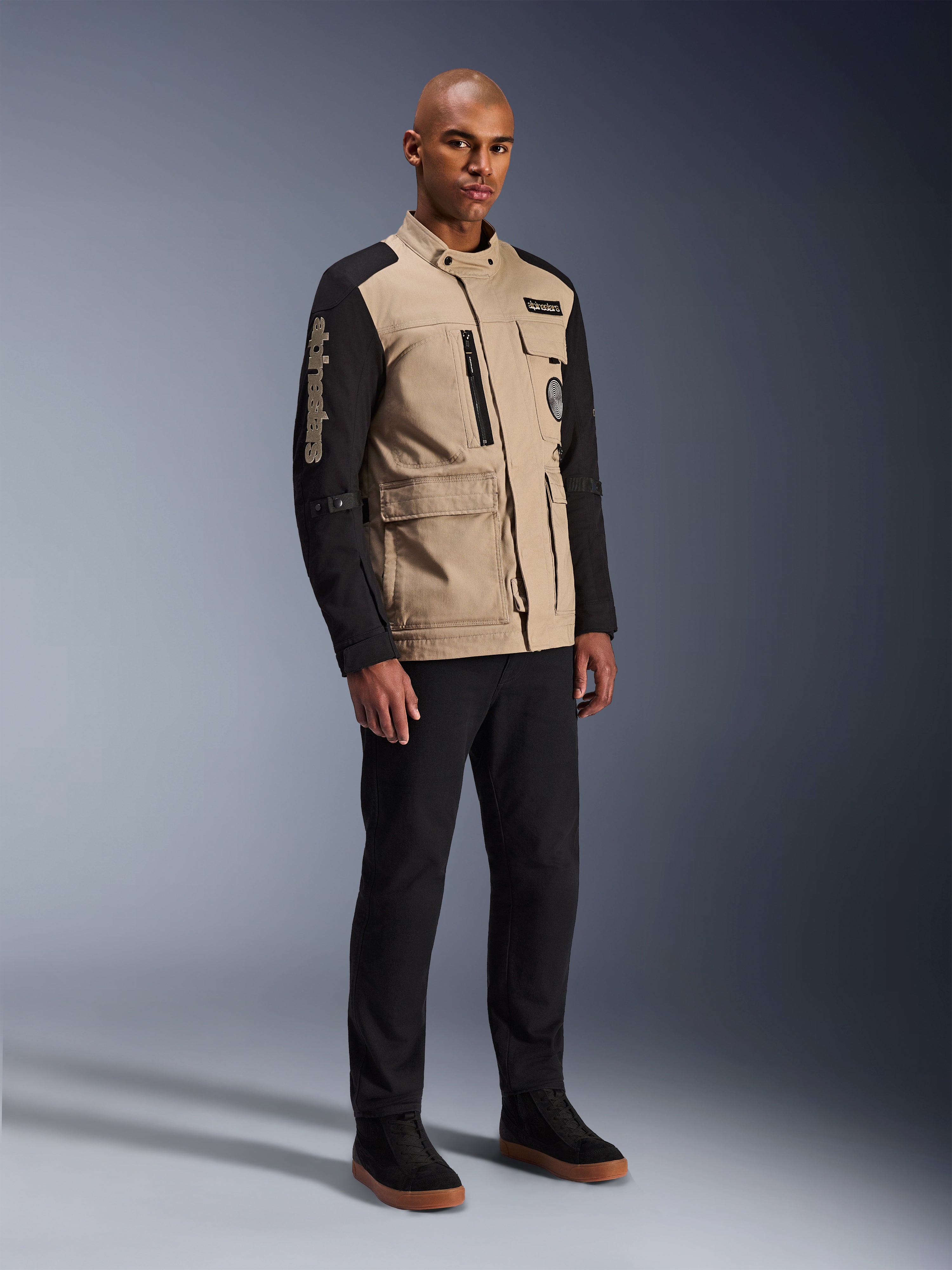 Veste Alpinestars Dice, veste de moto urbaine, Aluminum Black et Tan, design bicolore avec manches noires et buste kaki, portée par un mannequin avec un pantalon noir et des baskets noires, esthétique d'inspiration workwear avec poches utilitaires et marquage brodé sur la manche.