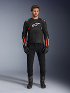 Männliches Model in der Alpinestars T-SPS Air v2 Jacke, schwarze Textil-Motorradjacke mit Red Fluo Akzenten, weißes Logo auf der Brust, getragen mit schwarzer Hose, Handschuhen und Stiefeln, urbaner sportlicher Fahrstil