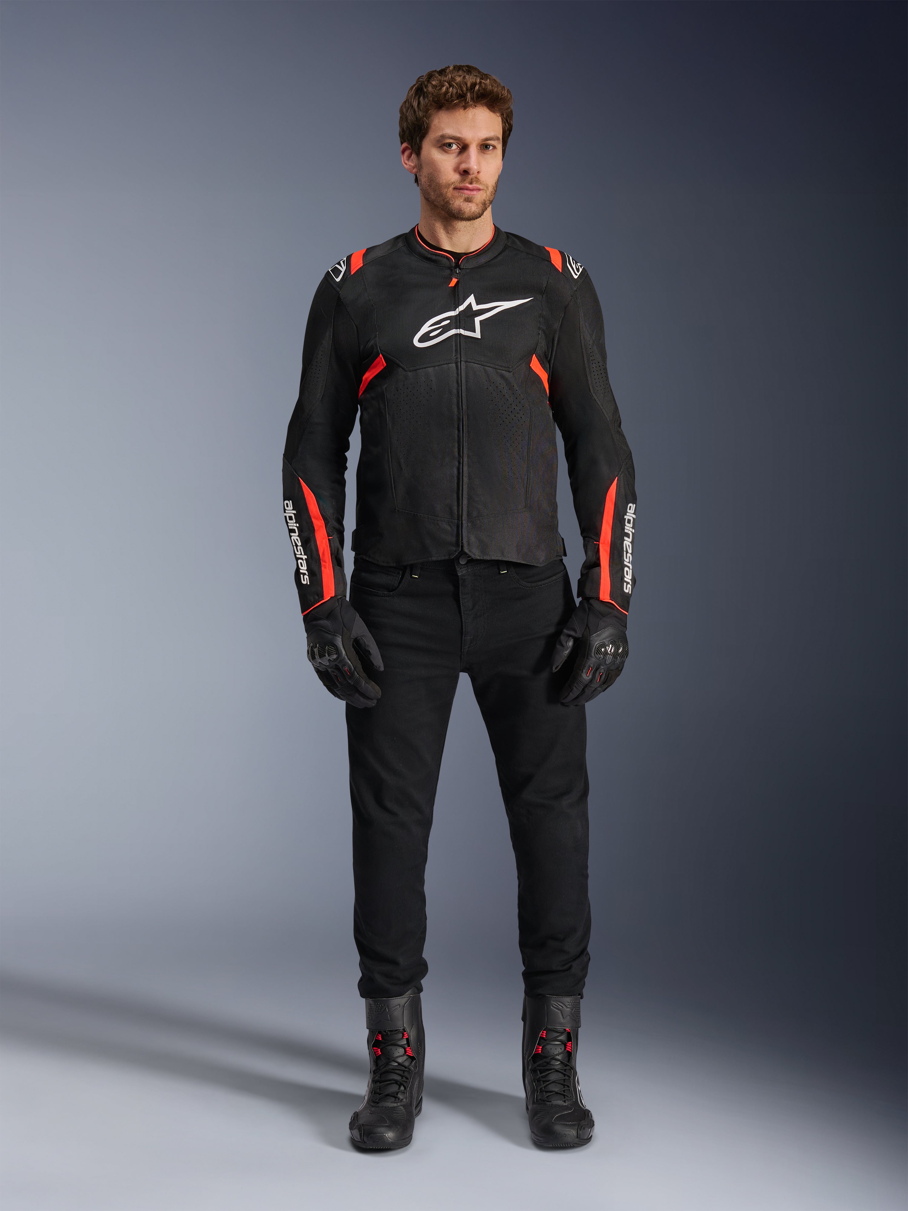 Männliches Model in der Alpinestars T-SPS Air v2 Jacke, schwarze Textil-Motorradjacke mit Red Fluo Akzenten, weißes Logo auf der Brust, getragen mit schwarzer Hose, Handschuhen und Stiefeln, urbaner sportlicher Fahrstil