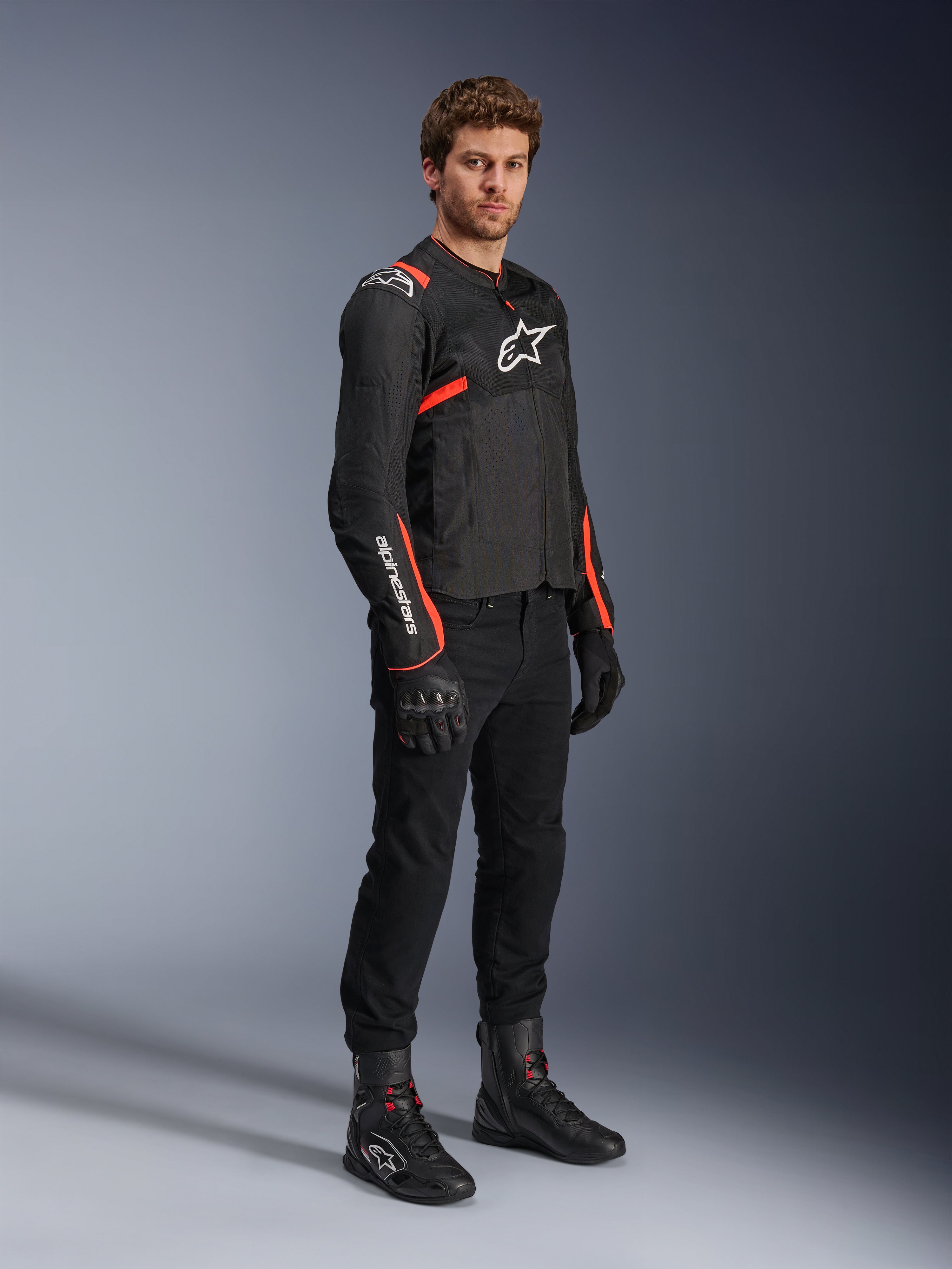 Männliches Model in der Alpinestars T-Sps Air V2 Jacke, Schwarz mit Red Fluo Akzenten, kombiniert mit schwarzer Motorradhose, schwarzen Handschuhen und schwarzen Stiefeln, sportliches Design mit Mesh-Einsätzen und weißen Alpinestars-Logos