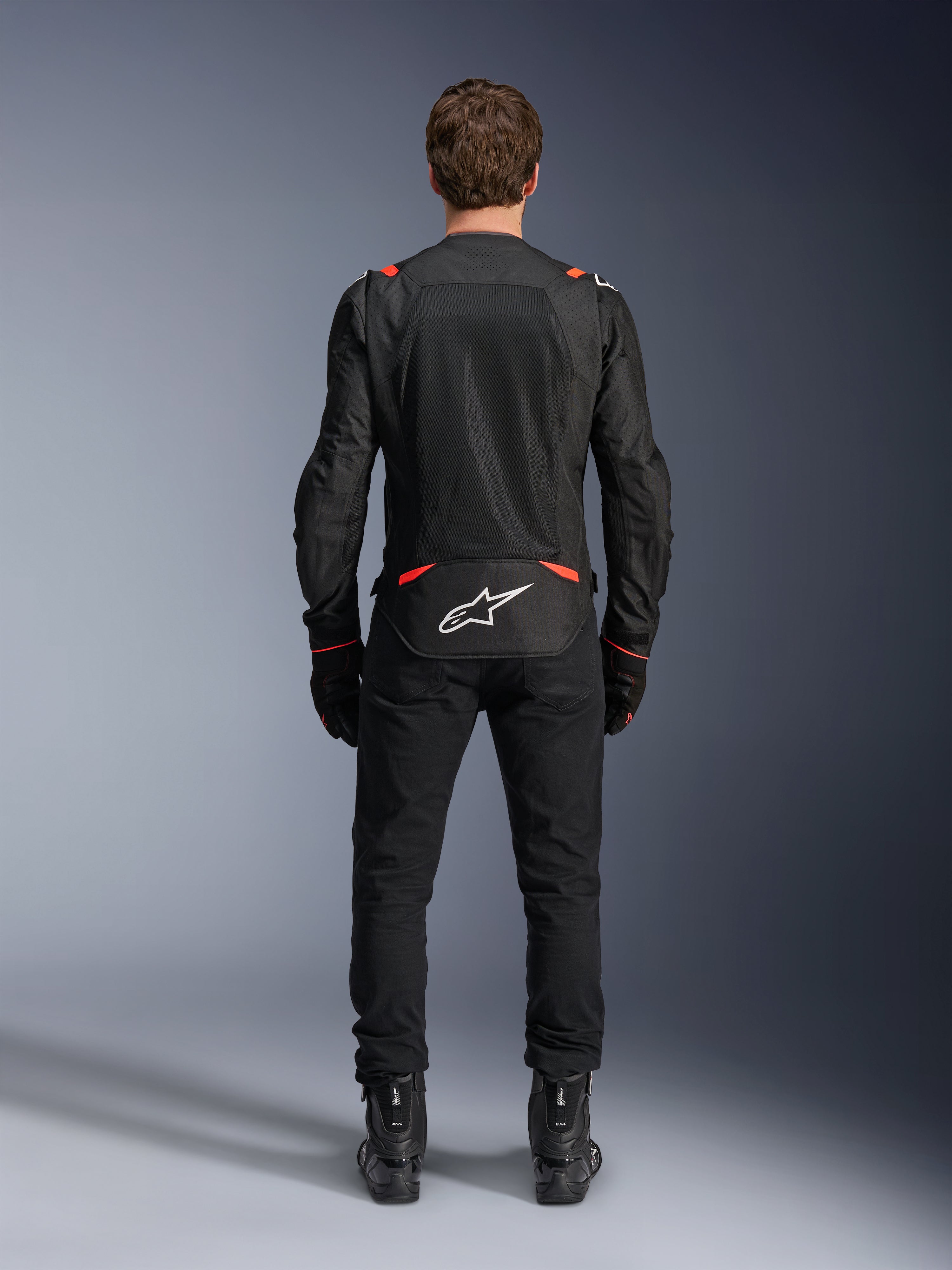 Alpinestars T-SPS Air v2 Jacke in Schwarz und Rot Fluo, Rückansicht mit großen Mesh-Belüftungseinsätzen und weißem Alpinestars-Logo auf dem unteren Rücken, getragen mit schwarzer Motorradhose und Stiefeln.