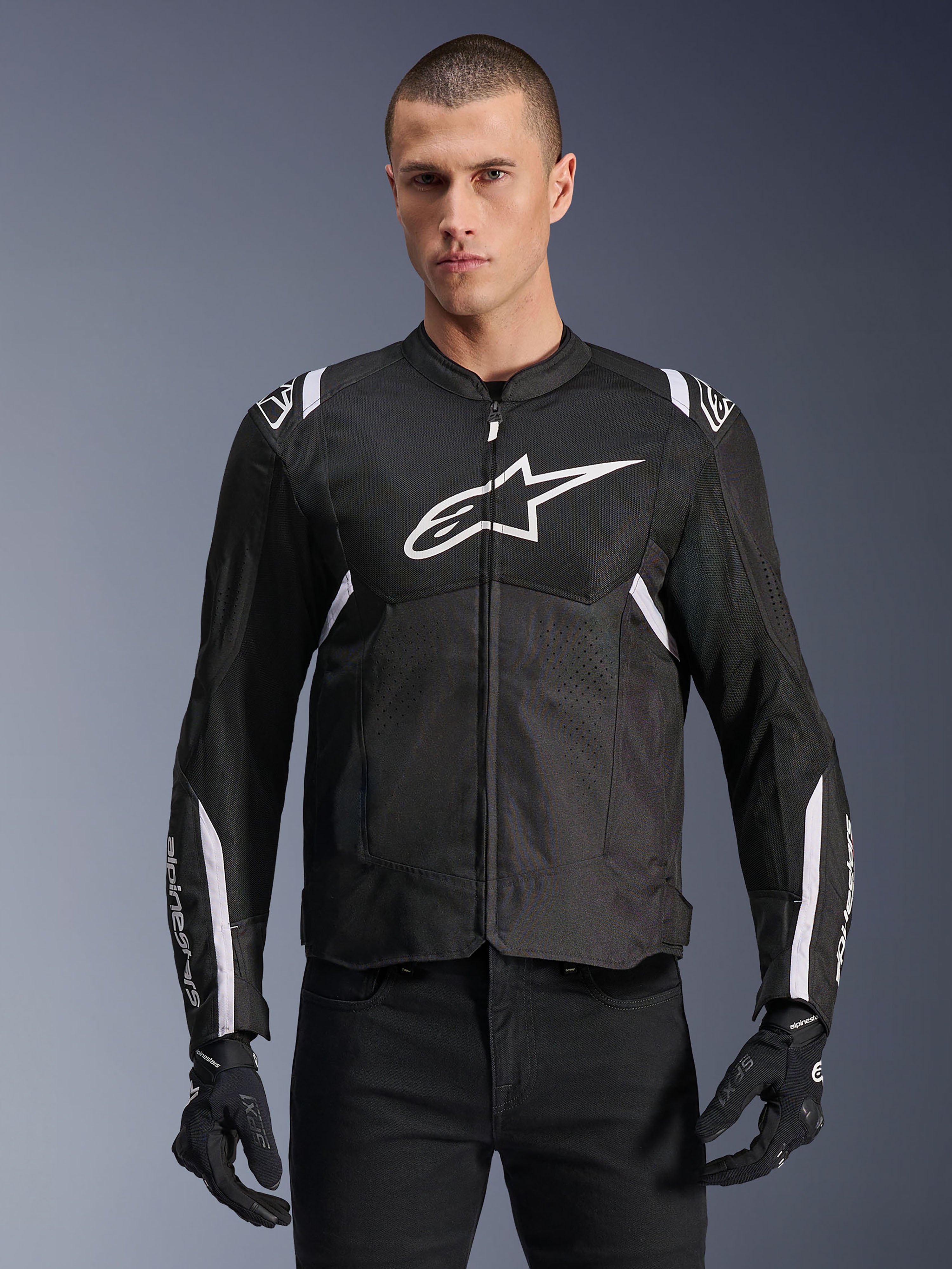 Alpinestars T-SPS Air V2 Jacke, Textil-Motorradjacke, Schwarz mit weißen Akzenten, großes weißes Brustlogo, perforiertes Mesh-Design für Belüftung, getragen mit schwarzer Hose und Handschuhen
