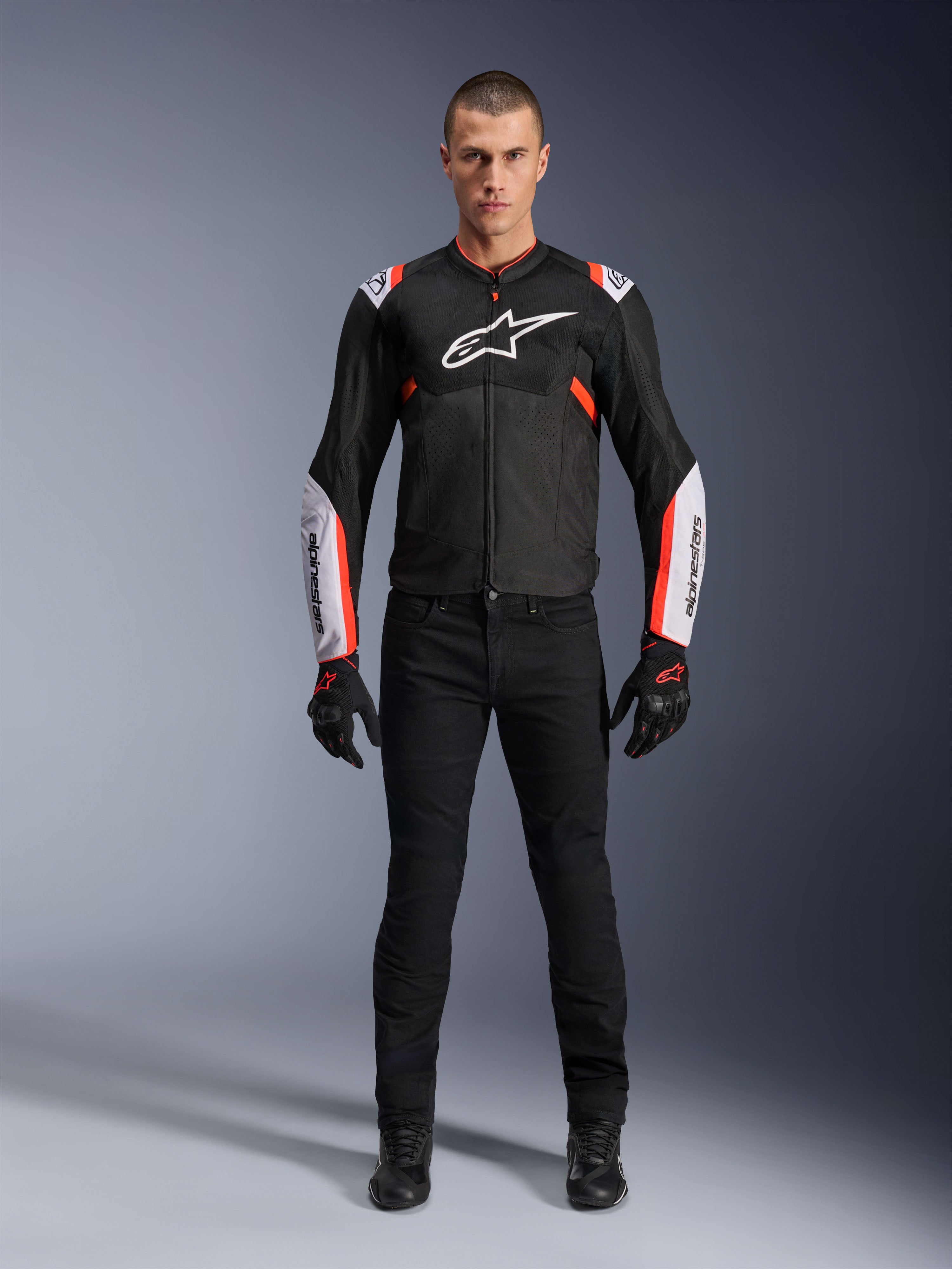 Männliches Model in der Alpinestars T-Sps Air V2 Jacke in Schwarz Weiß Rot Fluo, mit großen Tech-Mesh-Einsätzen und markantem weißem Branding auf Brust und Ärmeln, kombiniert mit schwarzer technischer Motorradhose und Handschuhen, urbane Sport-Motorradausrüstung