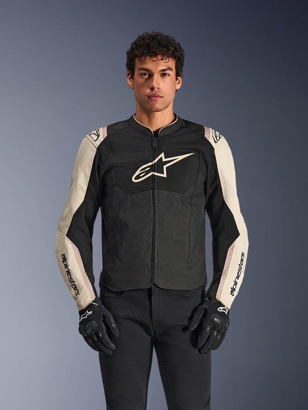 Veste Alpinestars T-SPS Air V2, veste de moto, noir et mastic aluminium, modèle masculin portant la veste avec un pantalon et des gants noirs, avec panneaux en maille pour la ventilation, grand logo Alpinestars sur la poitrine et design sportif ajusté