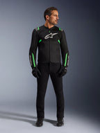 Ein Mann trägt die Alpinestars T-SPS Air V2 Jacke in Schwarz Fluo-Grün, kombiniert mit einer schwarzen Hose, Handschuhen und Motorradschuhen, mit sportlichem Schnitt, weißen Logos und Mesh-Einsätzen für einen modernen urbanen Look.