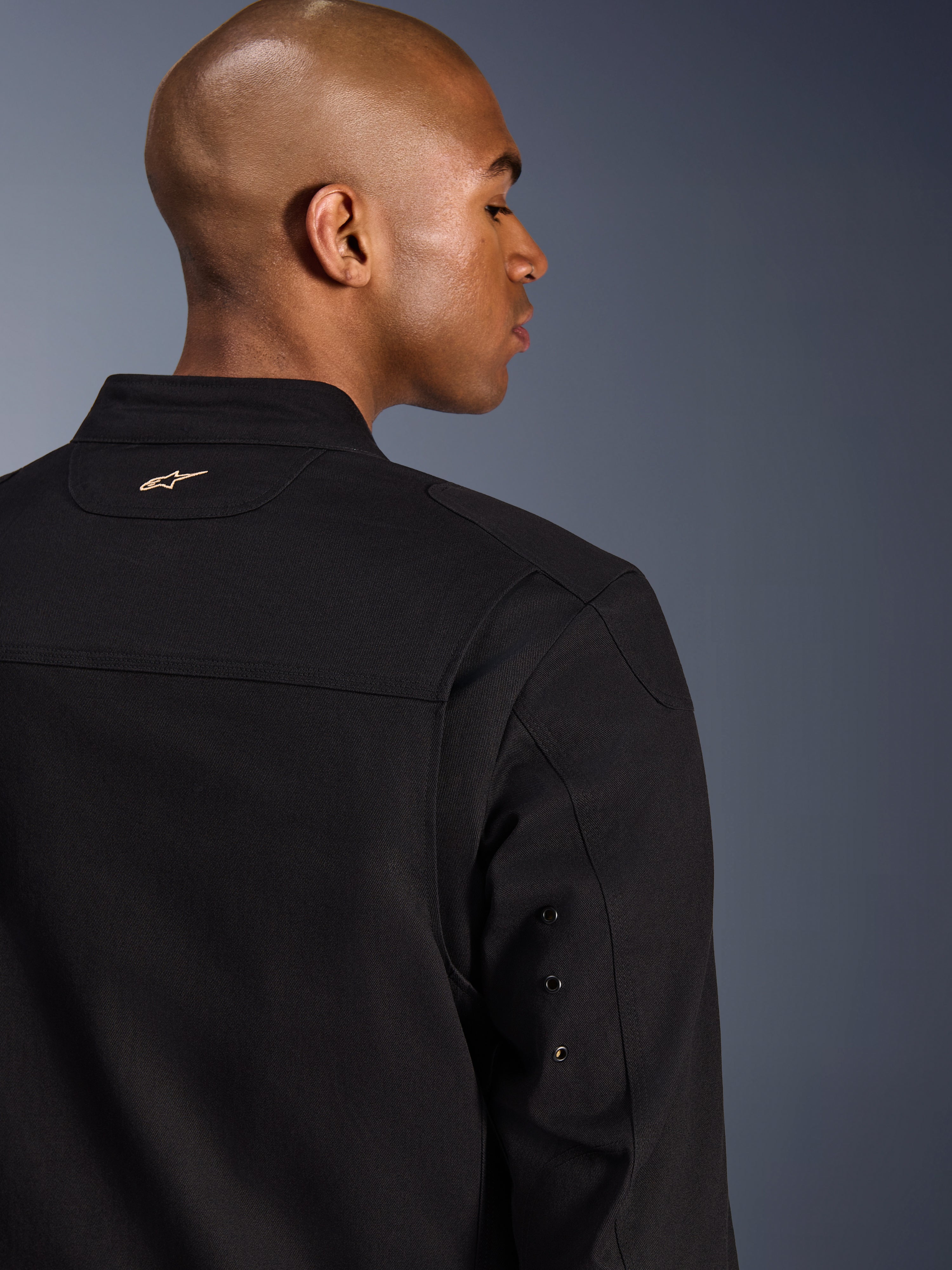 Alpinestars Bruiser Jacke, textile urbane Motorradjacke, Schwarz, Nahaufnahme der Rückansicht mit besticktem goldenem Logo am oberen Rücken, Metallösen-Details am Ärmel und plissierten Schulterbahnen für verbesserte Bewegungsfreiheit.