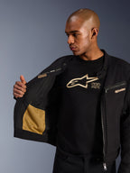 Alpinestars Bruiser Jacke, schwarze urbane Motorradjacke, Nahaufnahme der geöffneten Innenseite mit goldener Mesh-Tasche und „Alpinestars Protects“-Branding, getragen über einem schwarzen Logo-T-Shirt, textile Fahrerausrüstung mit Workwear-Ästhetik