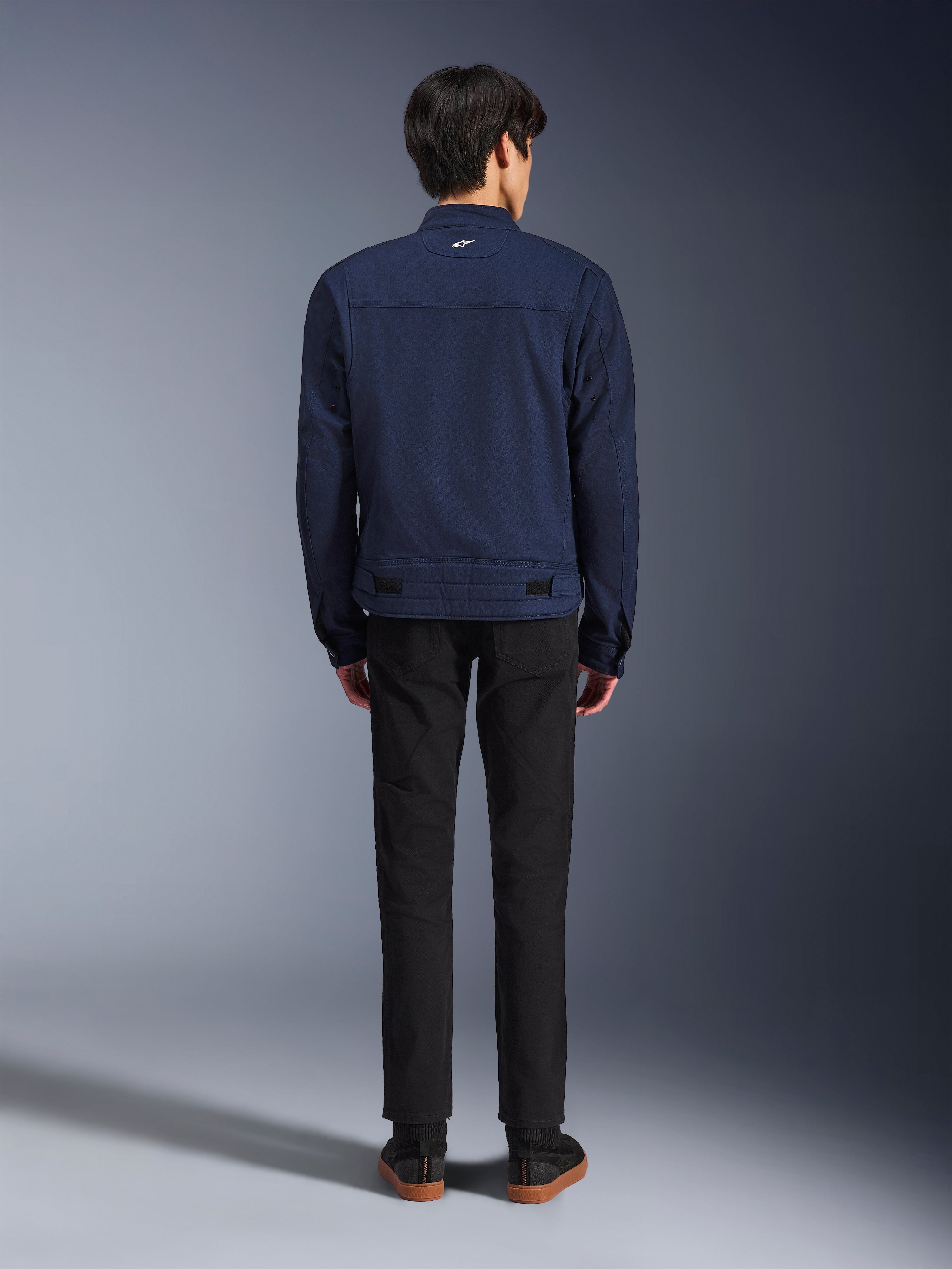 Veste Alpinestars Bruiser en bleu foncé, vue de dos sur mannequin, veste de moto en textile au design inspiré du vêtement de travail, dotée d'un petit logo blanc sur le col arrière et de pattes de réglage à la taille, associée à un pantalon noir