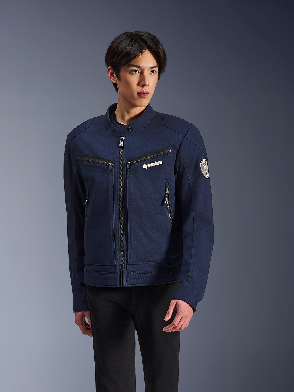 Veste Alpinestars Bruiser, veste moto en textile bleu foncé, portée par un homme avec un pantalon noir, avec poches zippées, logo Alpinestars blanc sur la poitrine et patch circulaire Oscar en spirale sur la manche, style streetwear urbain pour motards