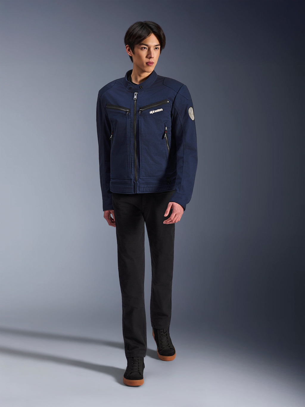 Modèle masculin portant la veste Alpinestars Bruiser en bleu foncé, veste de moto urbaine en textile, associée à un pantalon noir et des baskets noires, esthétique inspirée du vêtement de travail avec poches zippées et écusson circulaire sur la manche, streetwear contemporain pour la conduite à moto.