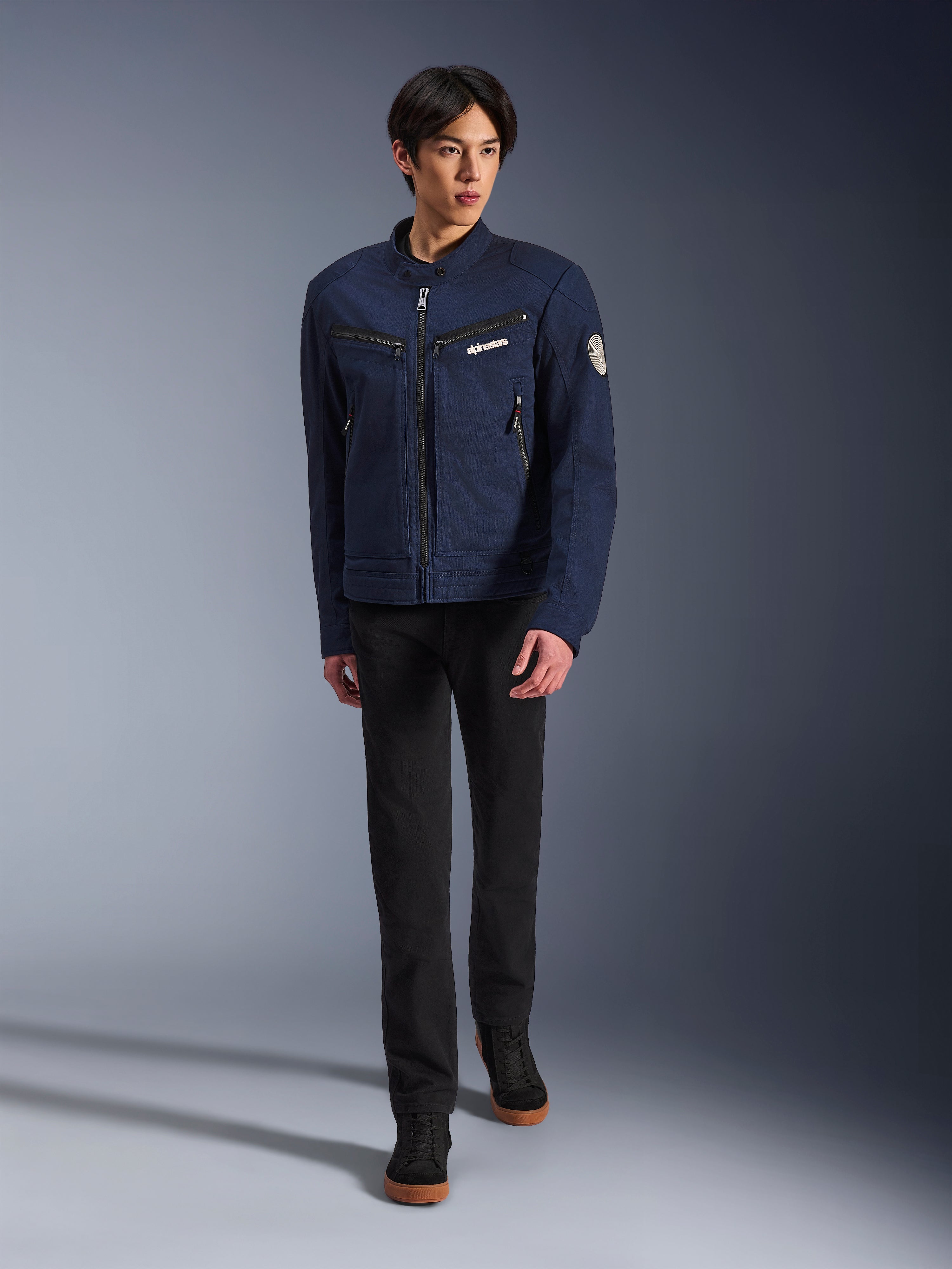 Modèle masculin portant la veste Alpinestars Bruiser en bleu foncé, veste de moto urbaine en textile, associée à un pantalon noir et des baskets noires, esthétique inspirée du vêtement de travail avec poches zippées et écusson circulaire sur la manche, streetwear contemporain pour la conduite à moto.