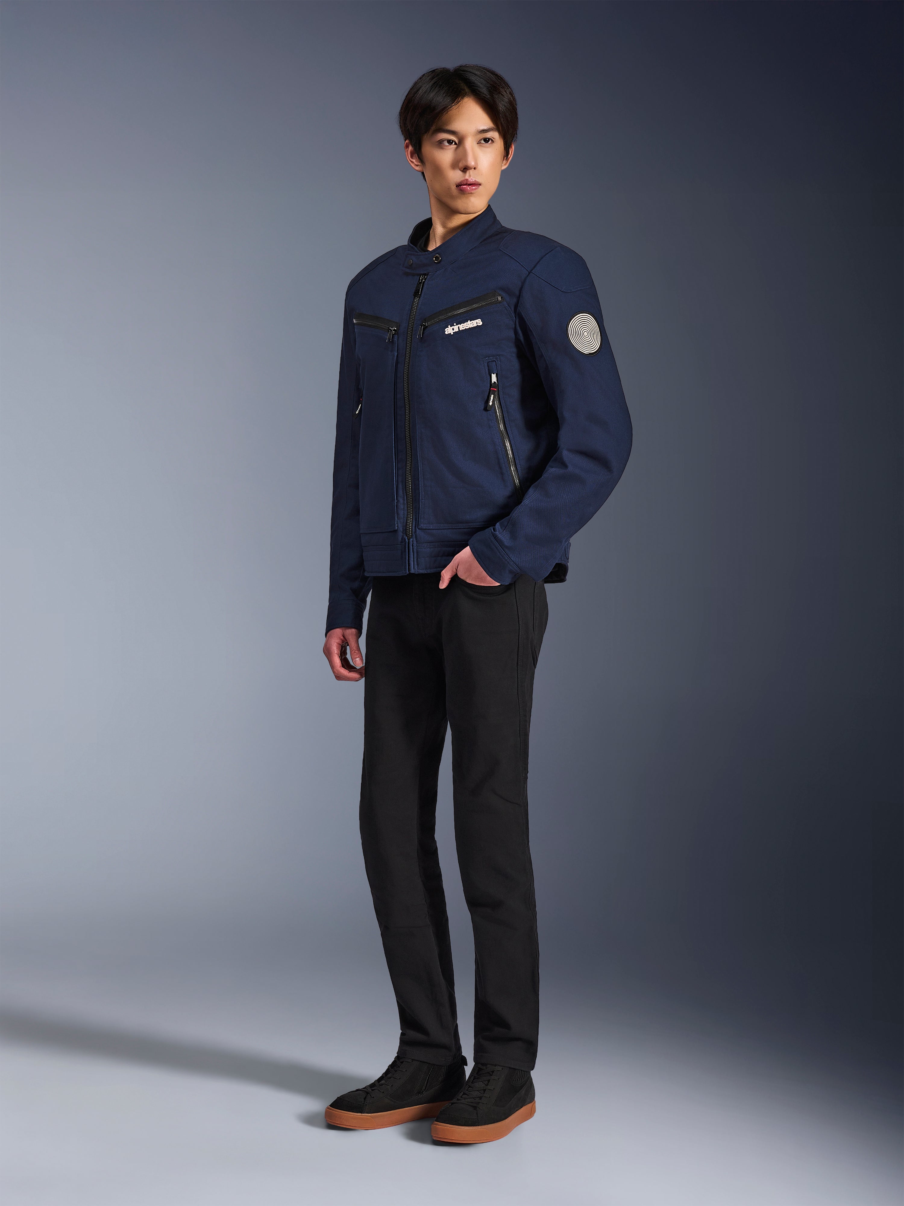 Veste Alpinestars Bruiser bleu foncé, veste de moto urbaine en textile au design inspiré du vêtement de travail, avec poches poitrine zippées et patch Oscar en spirale sur la manche, portée avec un pantalon noir et des baskets décontractées.