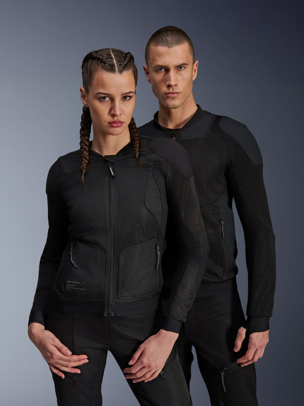 Alpinestars Troop-Air Pro Jas, zwarte technische gepantserde luchtjassen gedragen door een mannelijk en vrouwelijk model, voorzien van ademende mesh-panelen en geïntegreerde bescherming, urbane motorkleding ontworpen voor rijden in warm weer