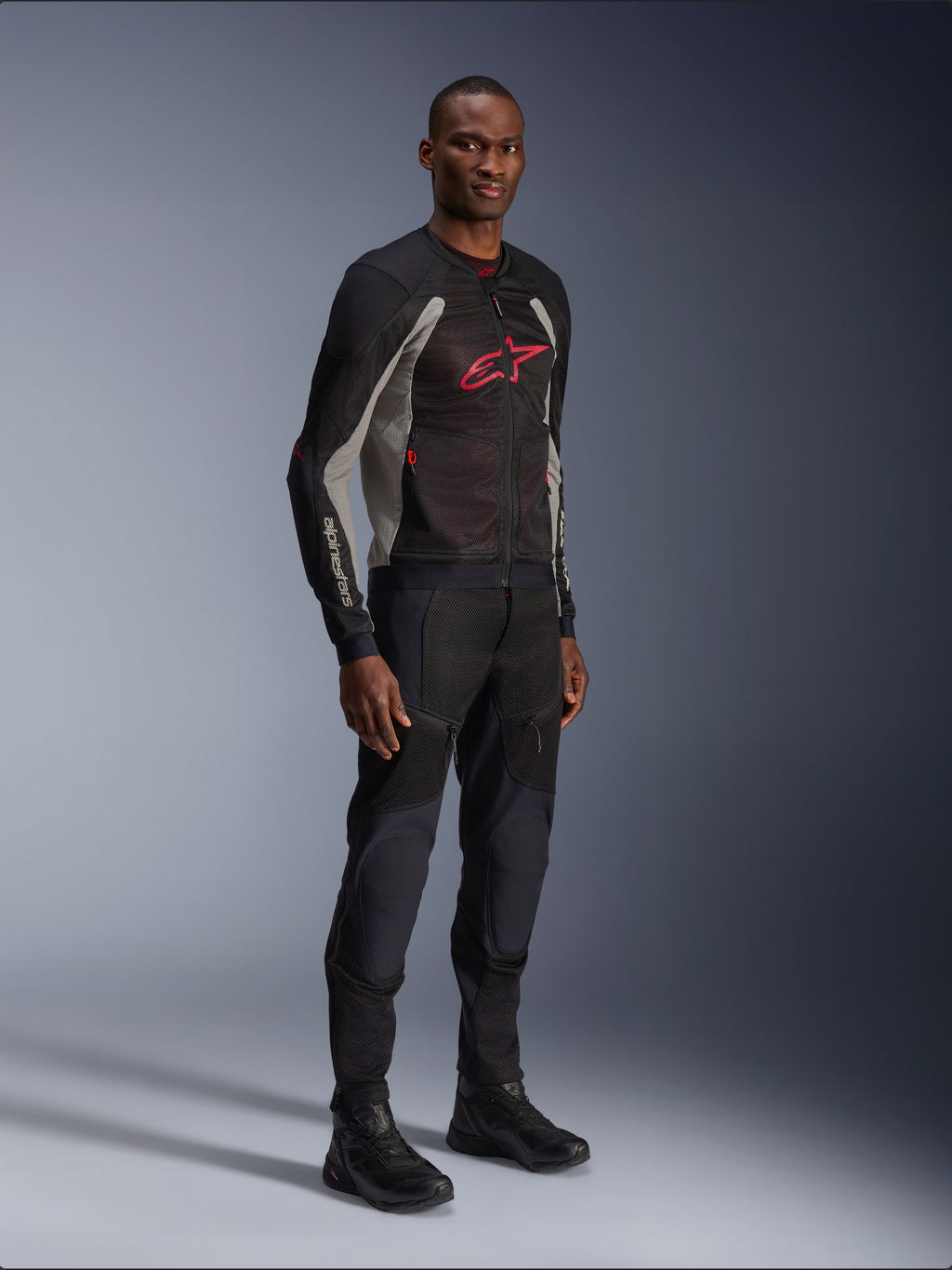 Alpinestars Troop-Air Jacke, technische Mesh-Motorradjacke in Schwarz, Ash Gray und Bright Red, getragen von einem Modell mit passender schwarzer Hose und Stiefeln, mit rotem Alpinestars-Logo auf der Brust, urbane zeitgemäße Motorradausrüstung.