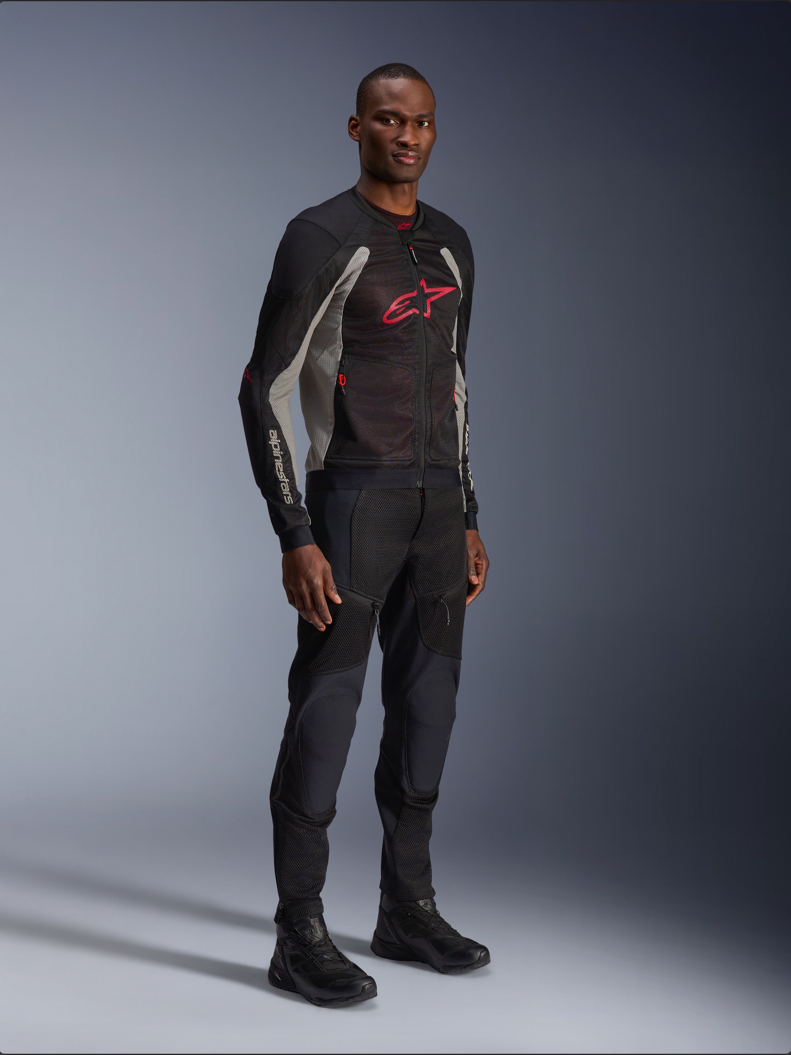 Alpinestars Troop-Air Jacke, technische Mesh-Motorradjacke in Schwarz, Ash Gray und Bright Red, getragen von einem Modell mit passender schwarzer Hose und Stiefeln, mit rotem Alpinestars-Logo auf der Brust, urbane zeitgemäße Motorradausrüstung.