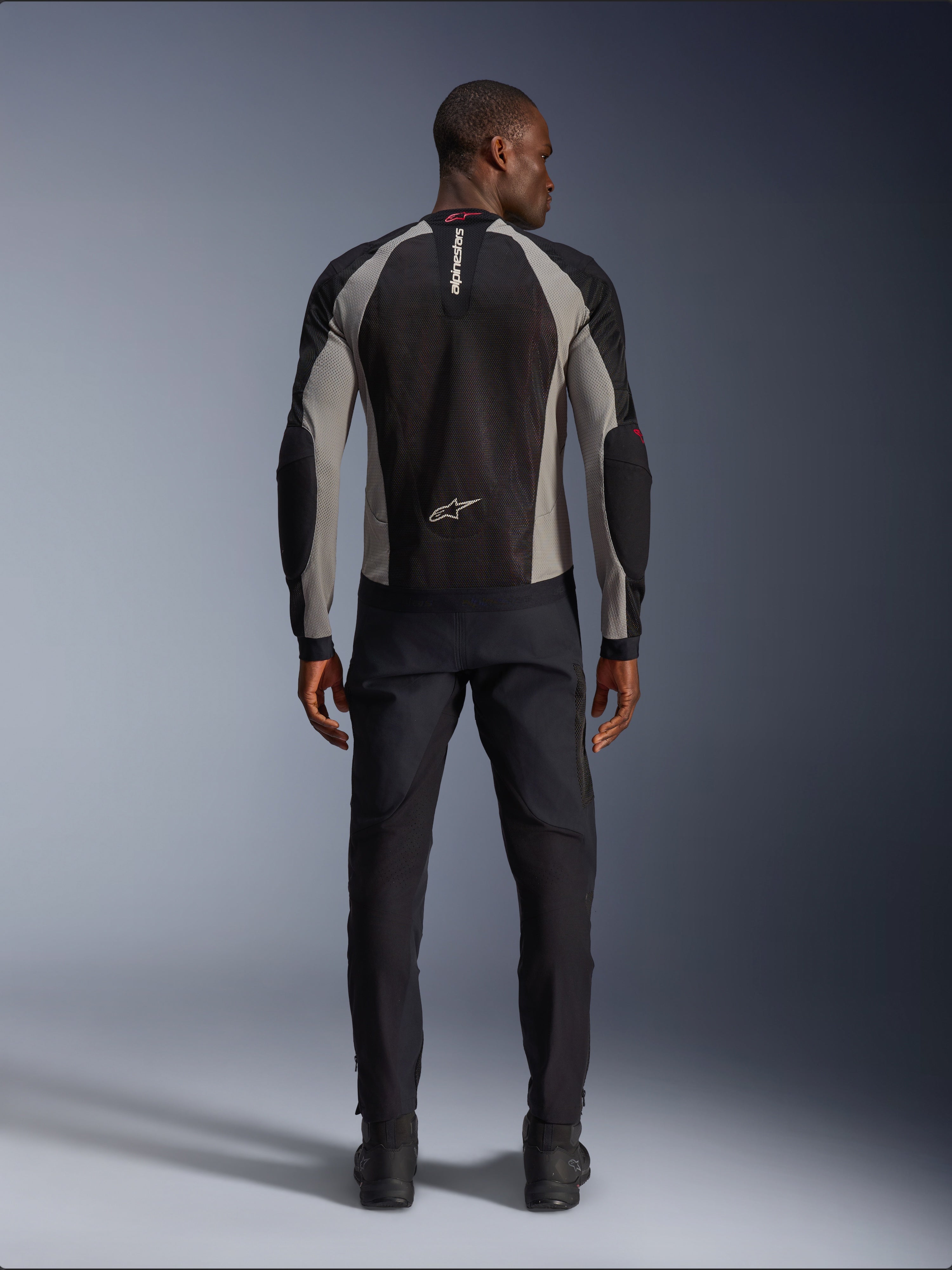 Alpinestars Troop-Air Jacke, Motorradjacke in Black Ash Gray und Bright Red, Rückansicht an einem männlichen Model, leichte Mesh-Konstruktion für Belüftung, Alpinestars-Branding am Kragen und im unteren Rückenbereich, kombiniert mit einer schwarzen Fahrhose.