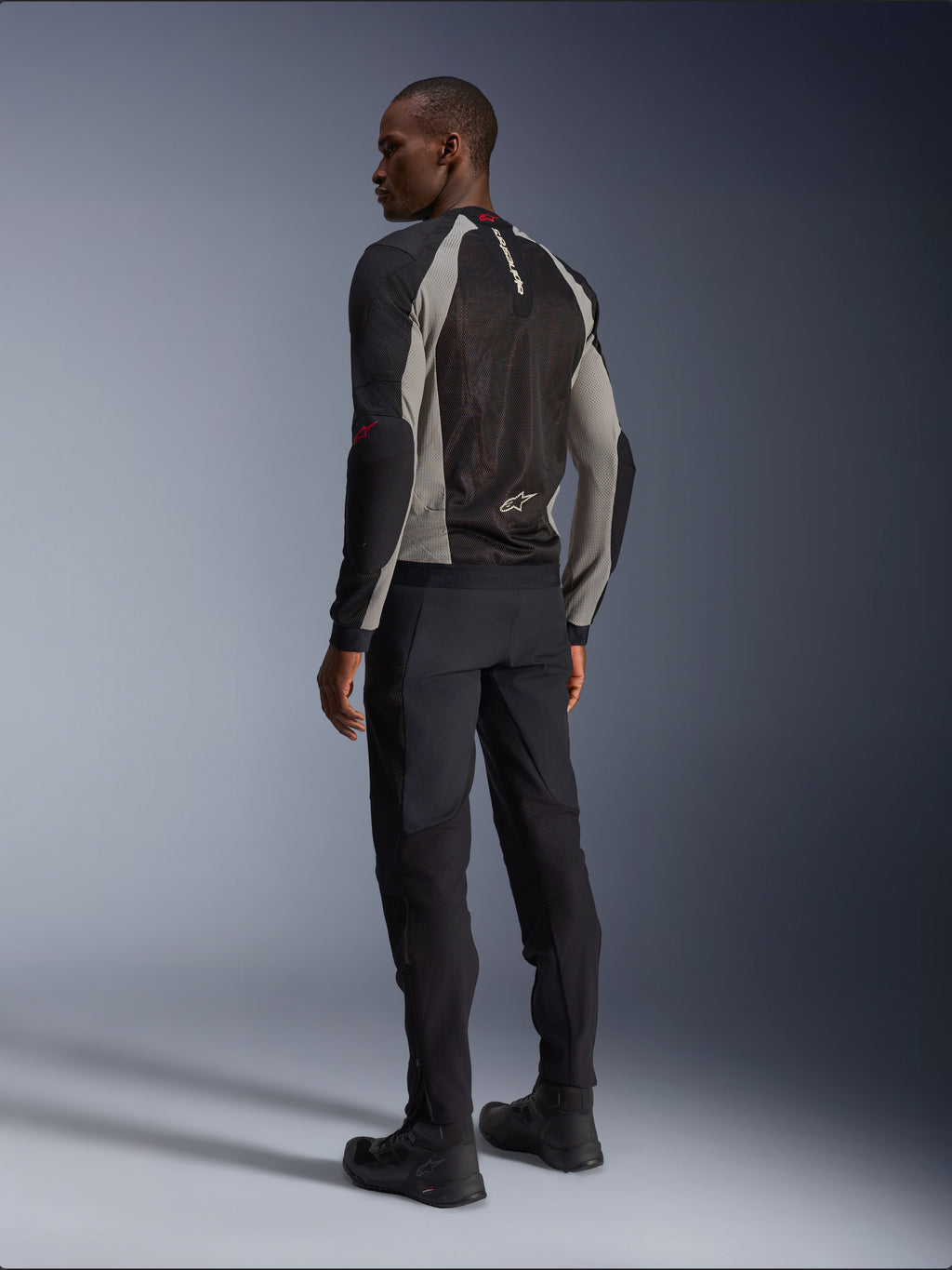 Model trägt Alpinestars Troop-Air Jacke in der Rückansicht, Schwarz, Aschgrau mit leuchtend roten Akzenten, technische Mesh-Konstruktion zur Belüftung, kombiniert mit schwarzer Motorradhose und Fahrstiefeln, urbaner zeitgenössischer Stil