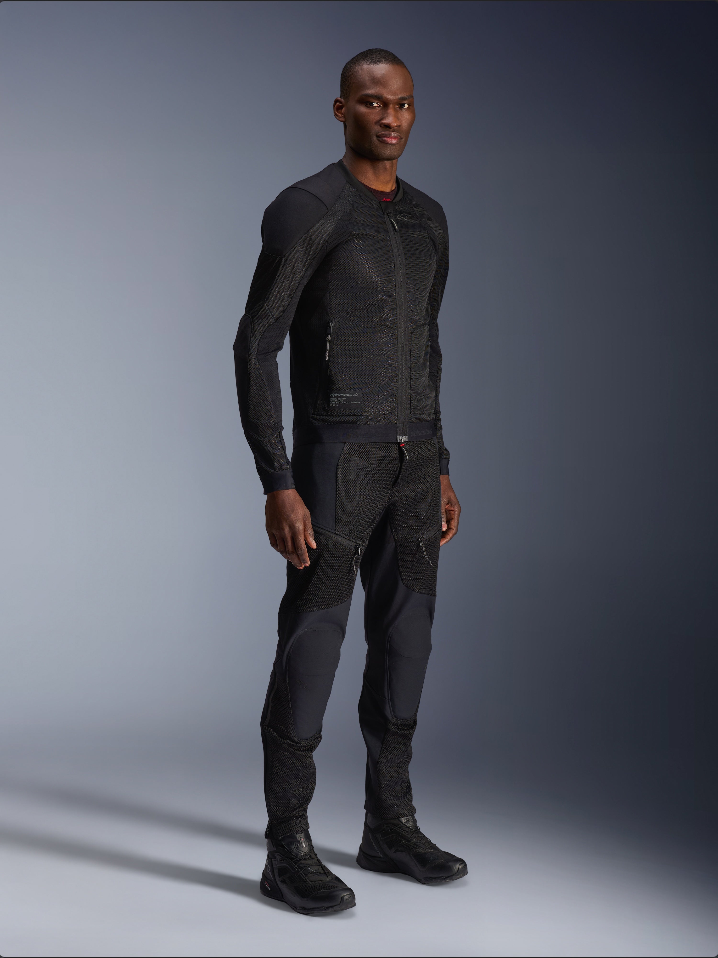 Alpinestars Troop-Air Jacket, zwart, technische mesh motorjas gedragen door een model met bijpassende zwarte broek en laarzen, met een lichtgewicht mesh-constructie en een minimalistisch urban design