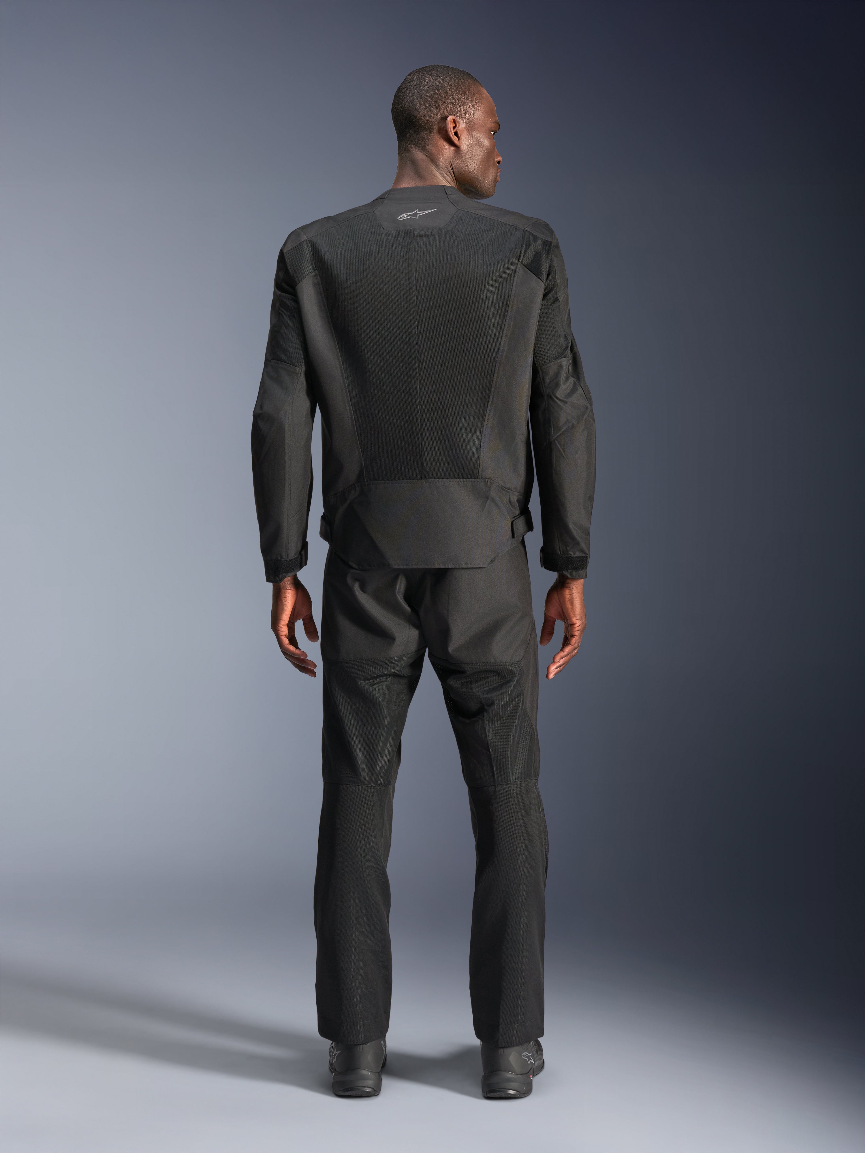 Rear view of a man wearing the Alpinestars C-1 Air Jacket in Black, urban motorcycle jacket with tech mesh panels, matching black textile pants and boots, clean aesthetic with reflective logo on the upper back.
