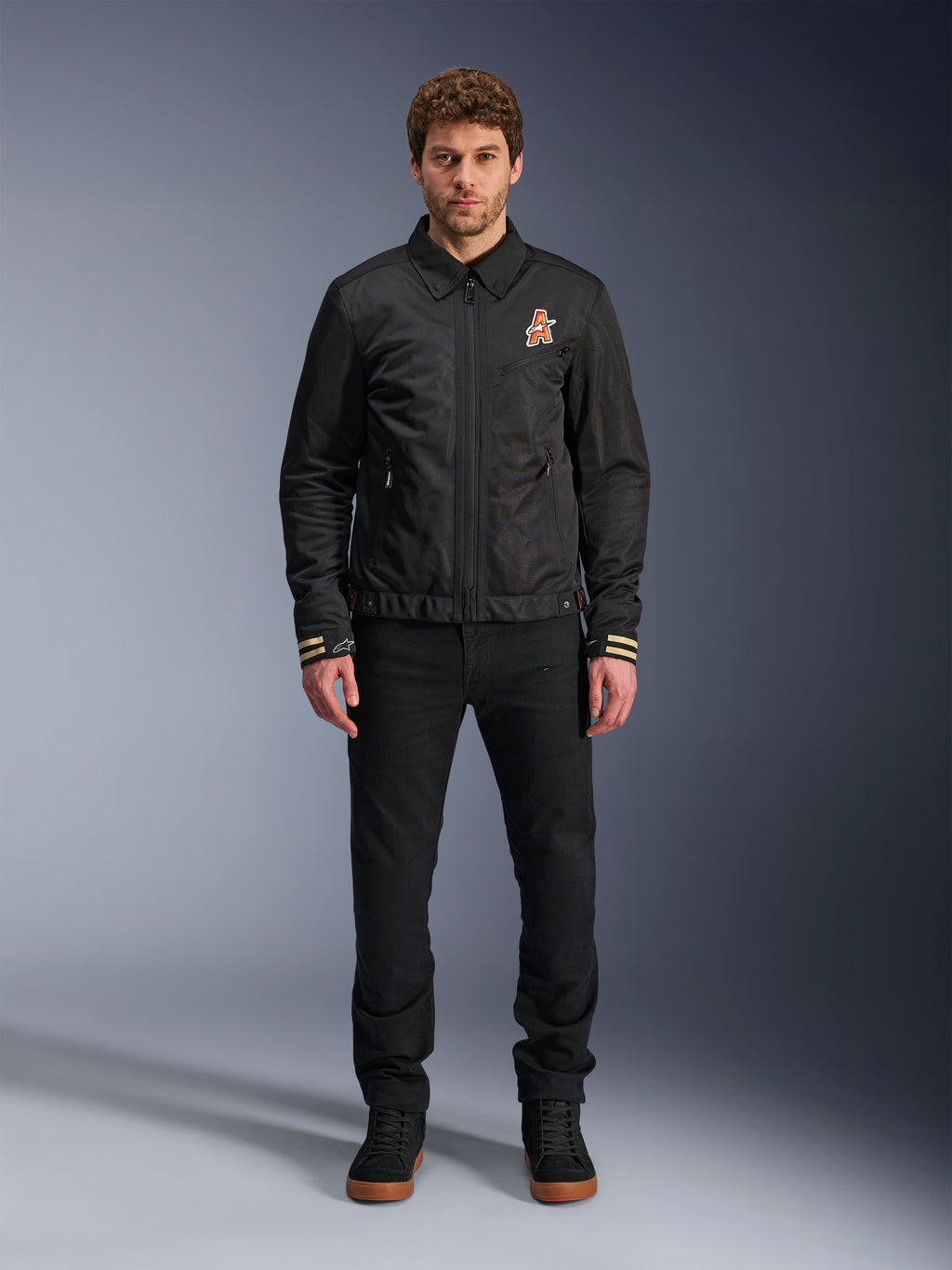 Männliches Model in der Alpinestars Ignite Air Jacke in Schwarz, mit klassischem Hemdkragen, Frontreißverschluss und einem orangefarbenen Logo auf der Brust. Kombiniert mit einer schwarzen Hose und schwarzen High-Top-Sneakern für einen modernen, urbanen Motorrad-Look.