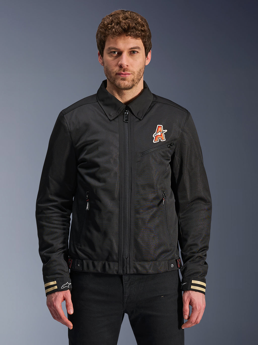 Alpinestars Ignite Air Jacket, schwarze Motorradjacke, traditioneller Hemdkragen, orange-weißes ‚A‘-Logo auf der Brust, beige gestreifte Manschetten mit weißem Stern-Logo, urbane, zeitgemäße Motorradausrüstung