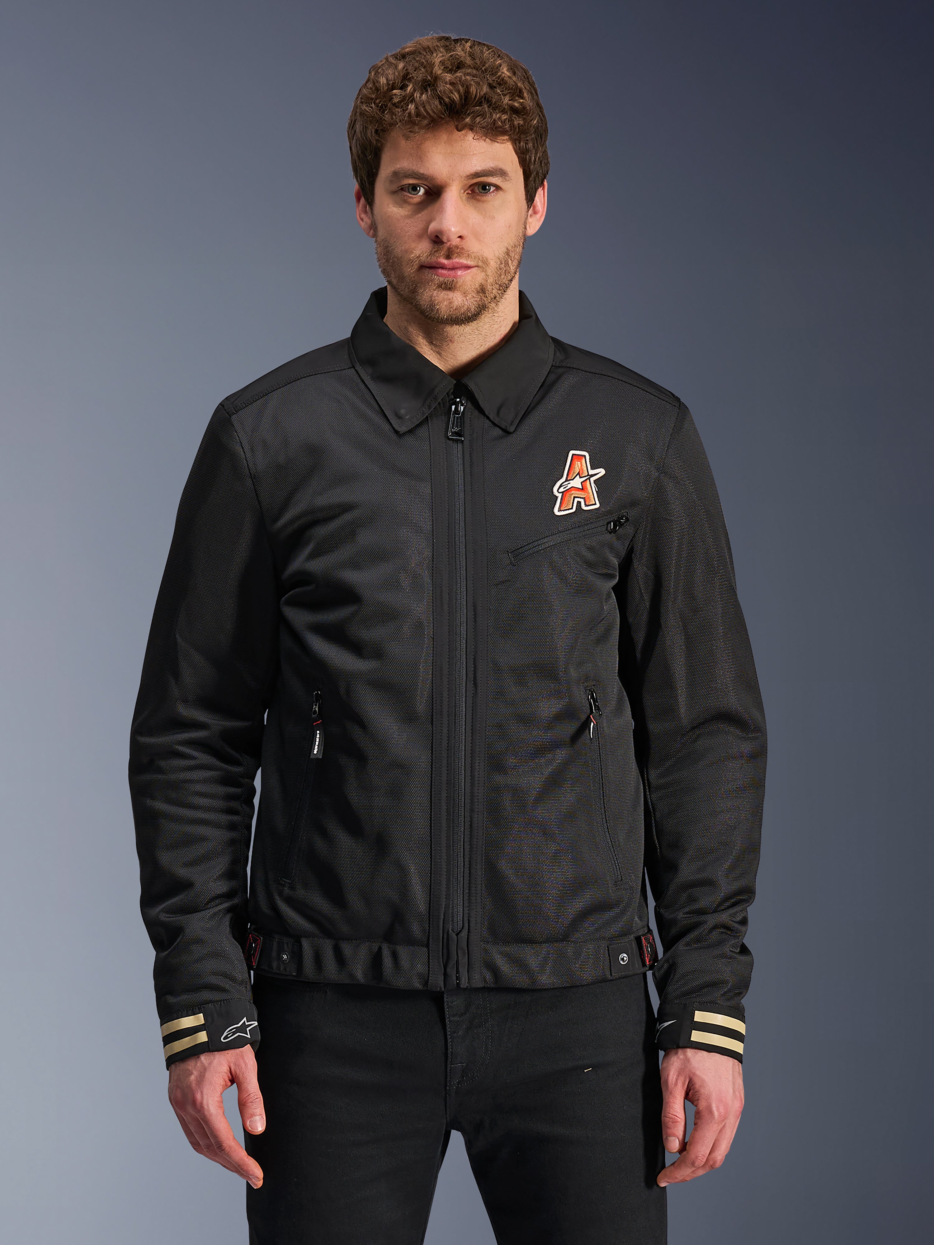 Alpinestars Ignite Air Jacket, schwarze Motorradjacke, traditioneller Hemdkragen, orange-weißes ‚A‘-Logo auf der Brust, beige gestreifte Manschetten mit weißem Stern-Logo, urbane, zeitgemäße Motorradausrüstung