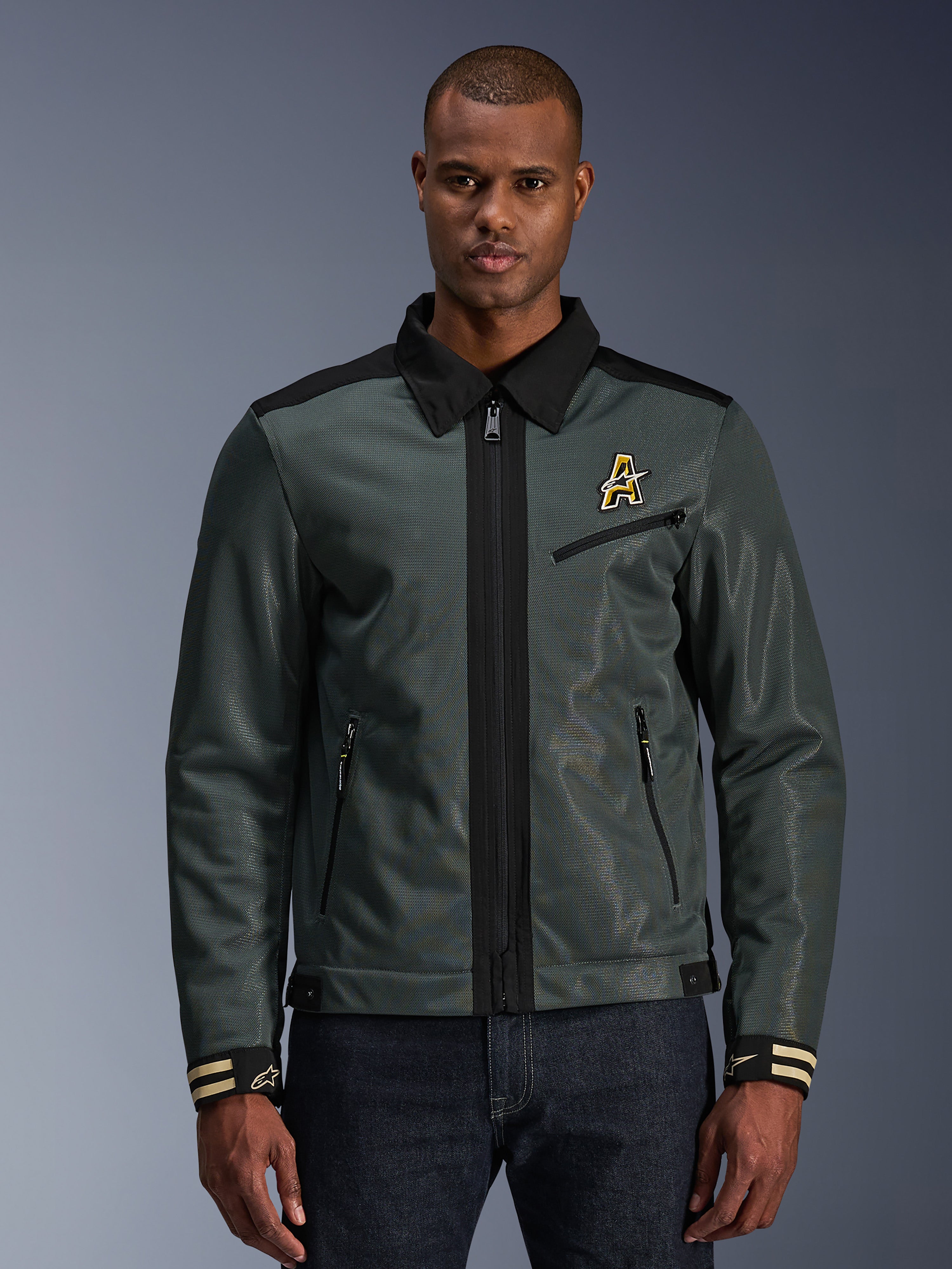 Alpinestars Ignite Air Jacket, urban motorjas, donker bosgroen met zwarte accenten, afgebeeld op model met denim broek, Harrington-stijl overhemdkraag, ritssluiting, logo op de borst en gestreepte manchetten, moderne mesh- en textielconstructie voor zomerse ritten