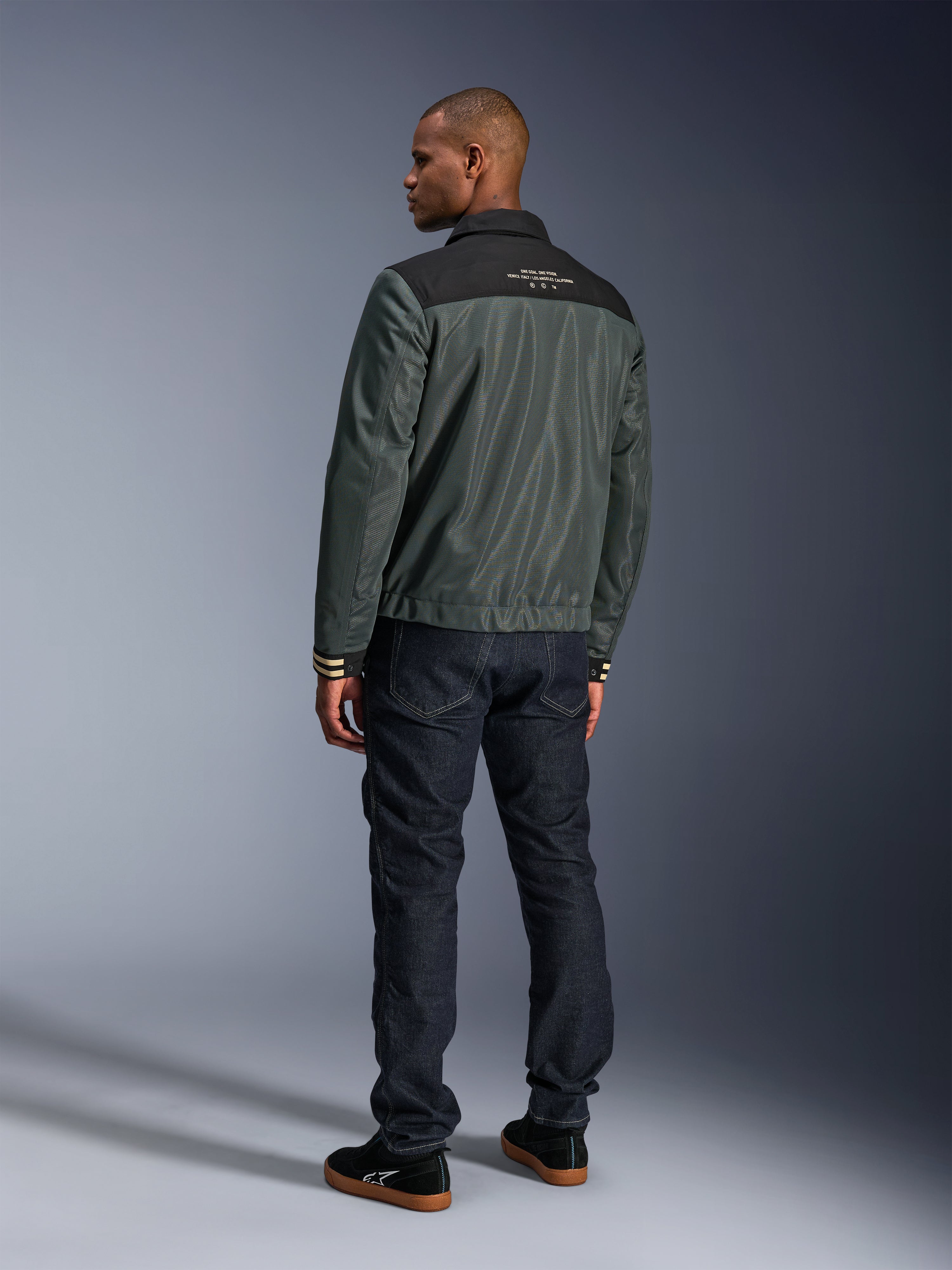 Veste Alpinestars Ignite Air en Gris Urbain Noir Bleu, vue de dos sur mannequin, dotée d'un corps en mesh avec un empiècement noir aux épaules et du texte blanc, poignets rayés, associée à un jean en denim foncé et des baskets noires, vêtement de moto style de vie urbain