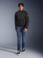 Männliches Model in der Alpinestars Provoke Jacke in Schwarz, urbane Textil-Motorradjacke mit technischen Reißverschlüssen und Ripstop-Softshell-Einsätzen, kombiniert mit blauen Jeans und schwarzen Sneakern, moderner urbaner Rider-Stil.