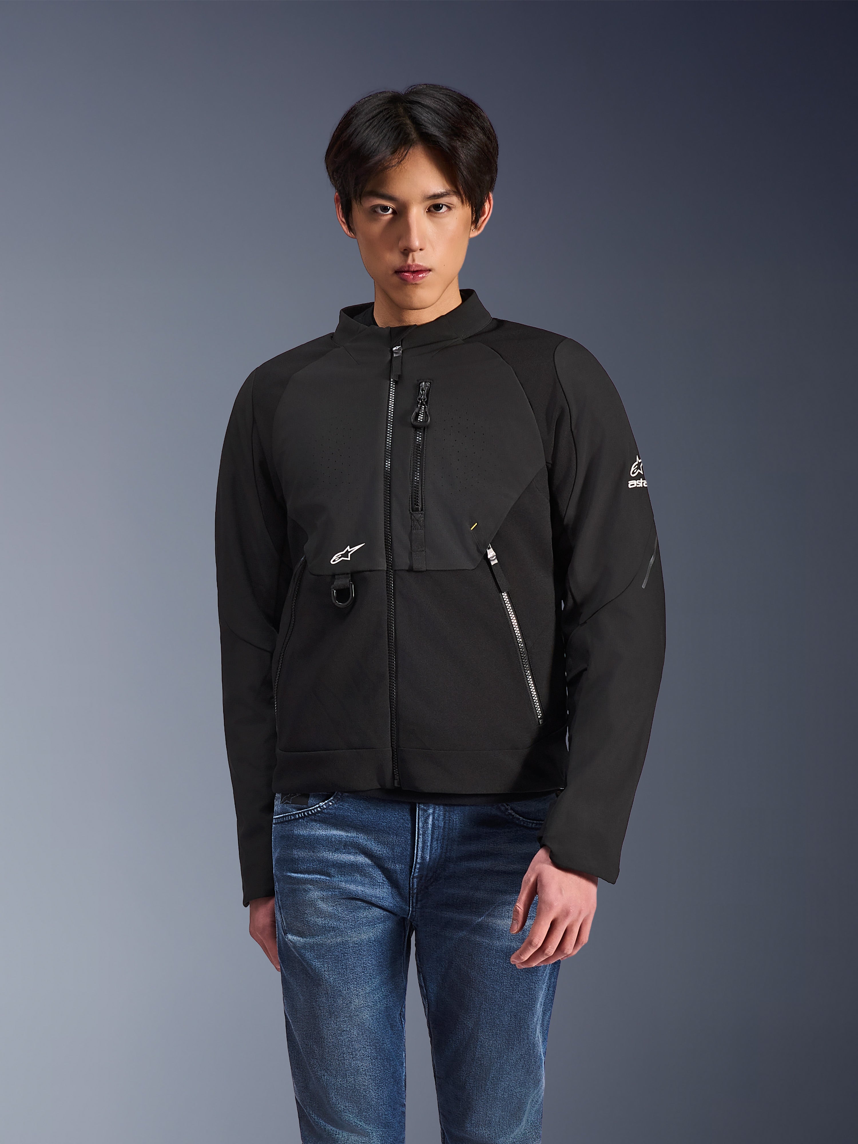 Veste Alpinestars Provoke, veste de moto urbaine en textile noir, portée par un mannequin homme avec un jean bleu, dotée de panneaux de poitrine perforés au laser, de plusieurs poches zippées, d'un logo A-star blanc sur la poitrine et la manche, design moderne et épuré pour la conduite urbaine