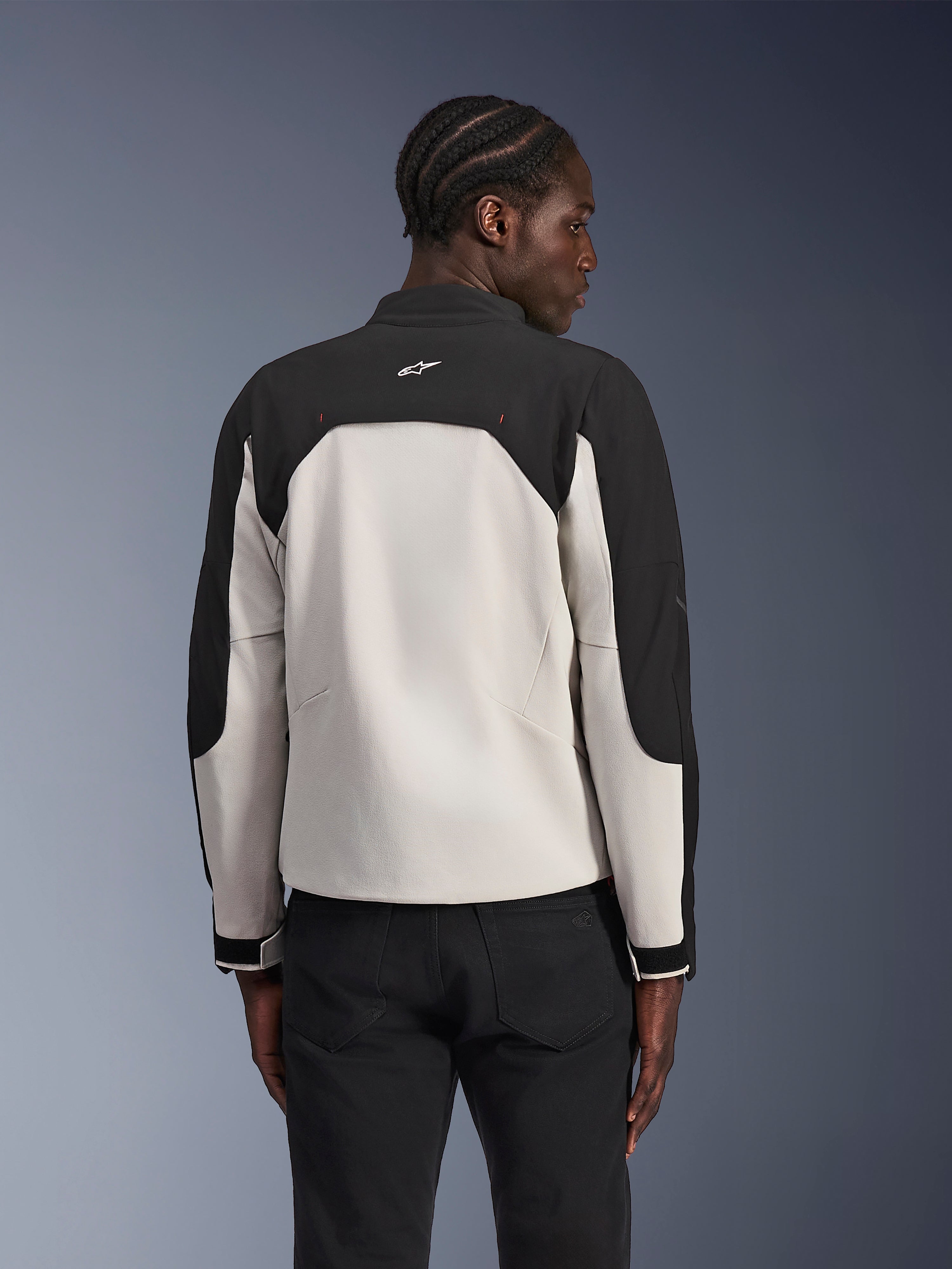 Veste Alpinestars Provoke, veste de moto urbaine, Noir Metal Gray, vue de dos sur un mannequin, présentant un design textile bicolore avec des épaules noires et un panneau arrière gris clair, logo étoile Alpinestars sur le col arrière, associée à un pantalon noir sur un fond dégradé bleu