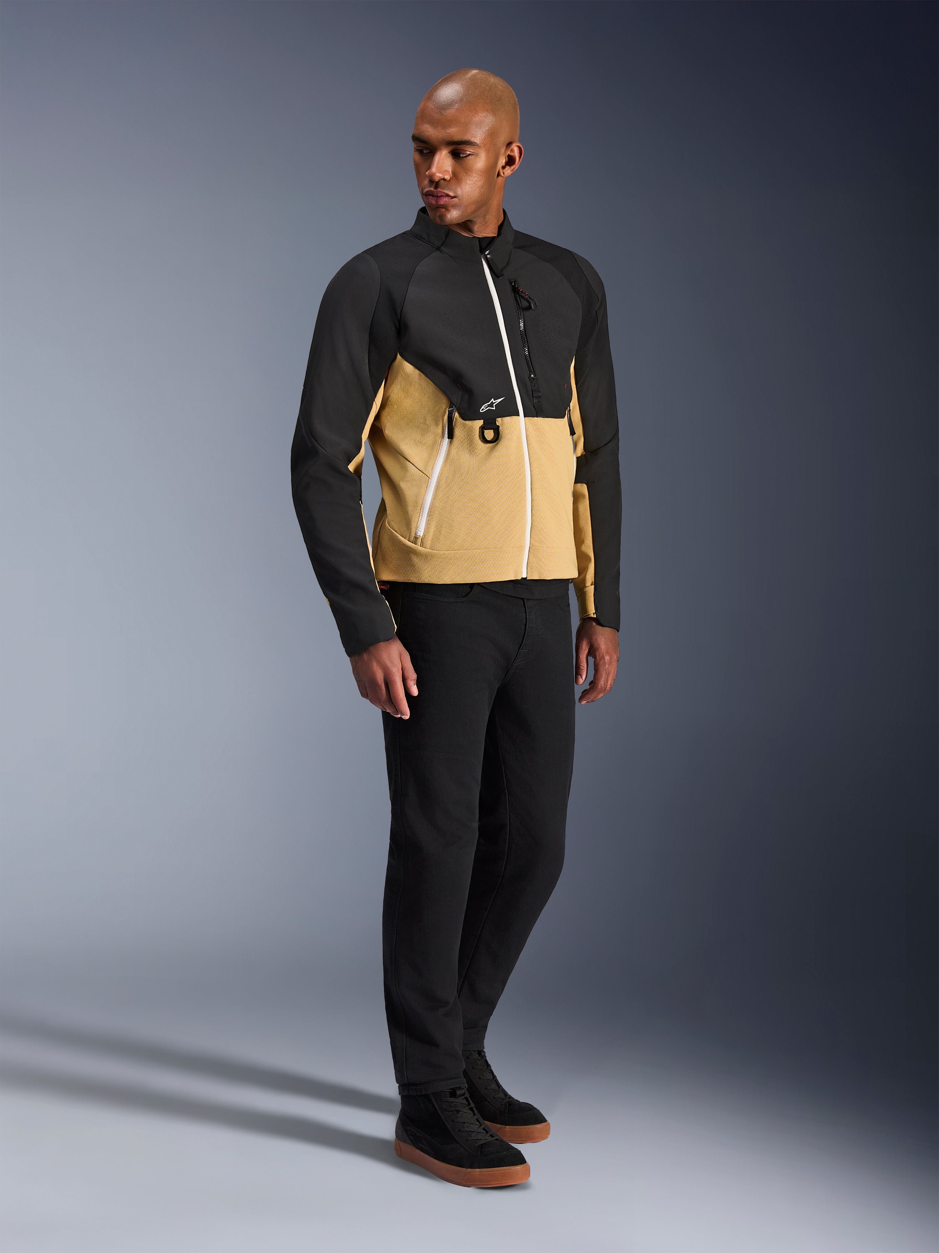 Veste Alpinestars Provoke, veste de moto urbaine, Black Camel, portée par un mannequin avec un pantalon noir et des baskets, design textile bicolore avec le haut noir et le bas du buste camel, détails de fermetures éclair blanches, équipement de moto urbain contemporain