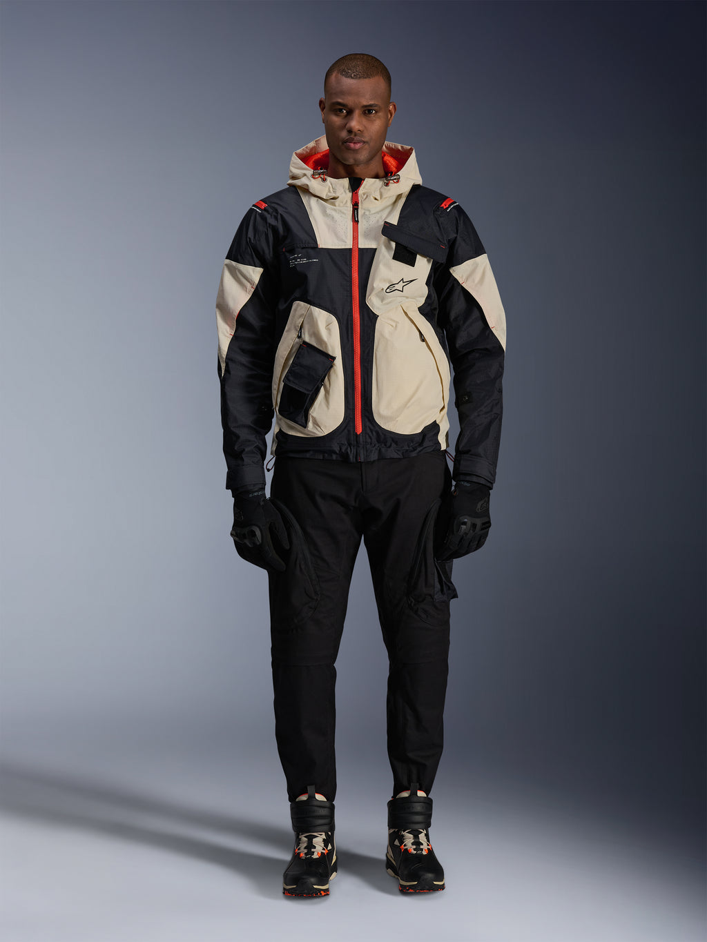 Alpinestars Mogress Air Jacket, Urban Motorcycle Jacket, Black Almond and Hot Orange, worn by a male model with black pants and gloves, utility-inspired design featuring cream panels and bright orange zippers, modern motorsport-influenced streetwear.