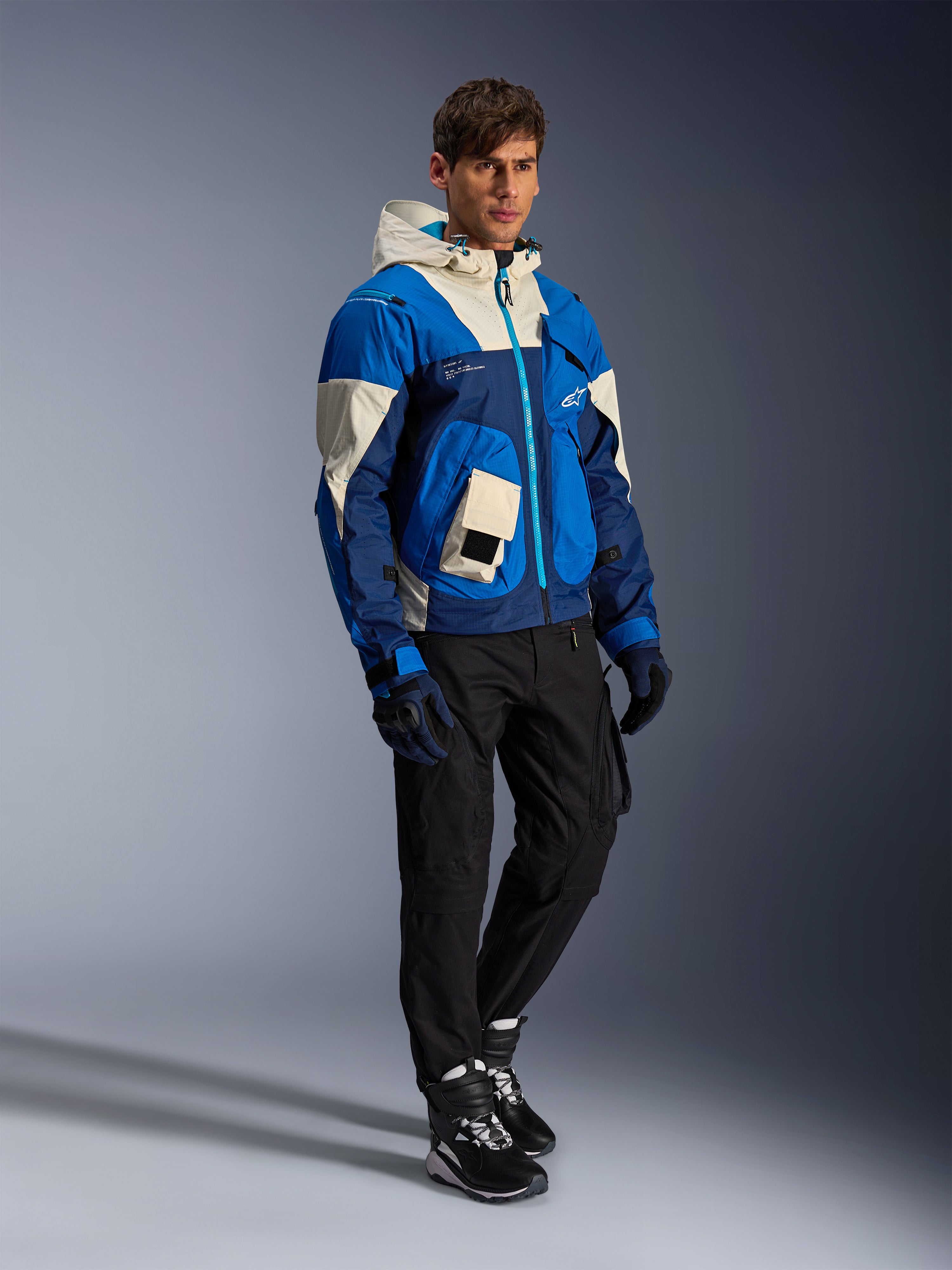 Alpinestars Mogress Air Jacke, urbane Motorradjacke, Blue Almond mit cremefarbenen und marineblauen Einsätzen, mit Kapuze und Utility-Taschen, getragen mit schwarzer Motorradhose, Handschuhen und Stiefeln, moderne, von Streetwear inspirierte Schutzausrüstung.