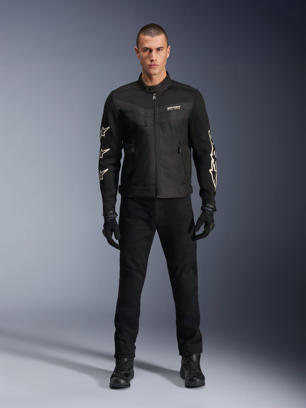 Modèle masculin portant la veste Alpinestars T-Dyno Air, noire, veste de moto légère en textile avec maille 3D respirante, logos d'étoiles blanches sur les manches, avec pantalon, gants et bottes noirs, vue de face debout