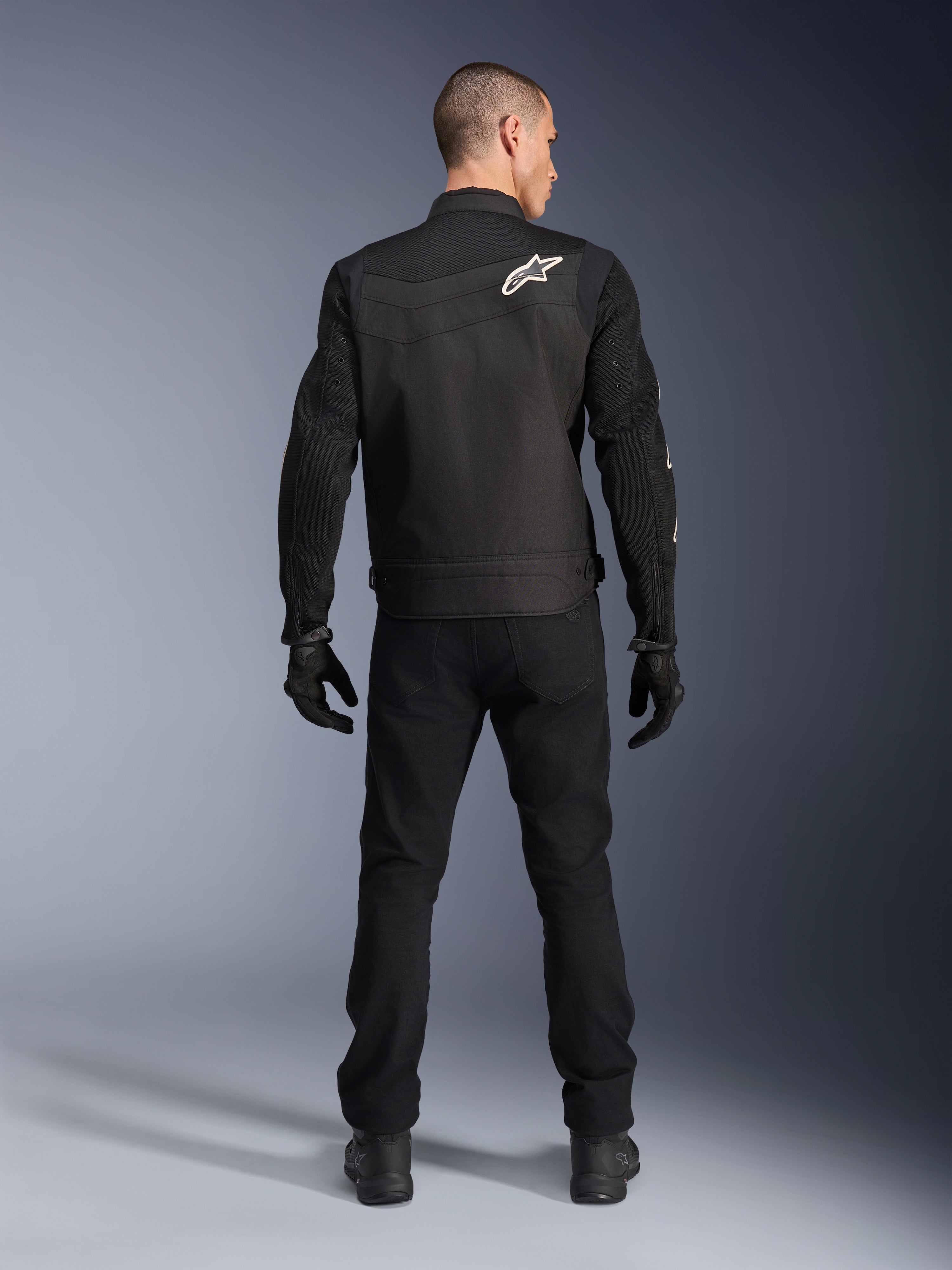 Veste Alpinestars T-Dyno Air, noire, vue arrière sur modèle, veste de moto textile dotée de panneaux en maille 3D et d'un logo étoile blanc sur l'épaule, portée avec un pantalon et des gants noirs, tenue de conduite urbaine moderne