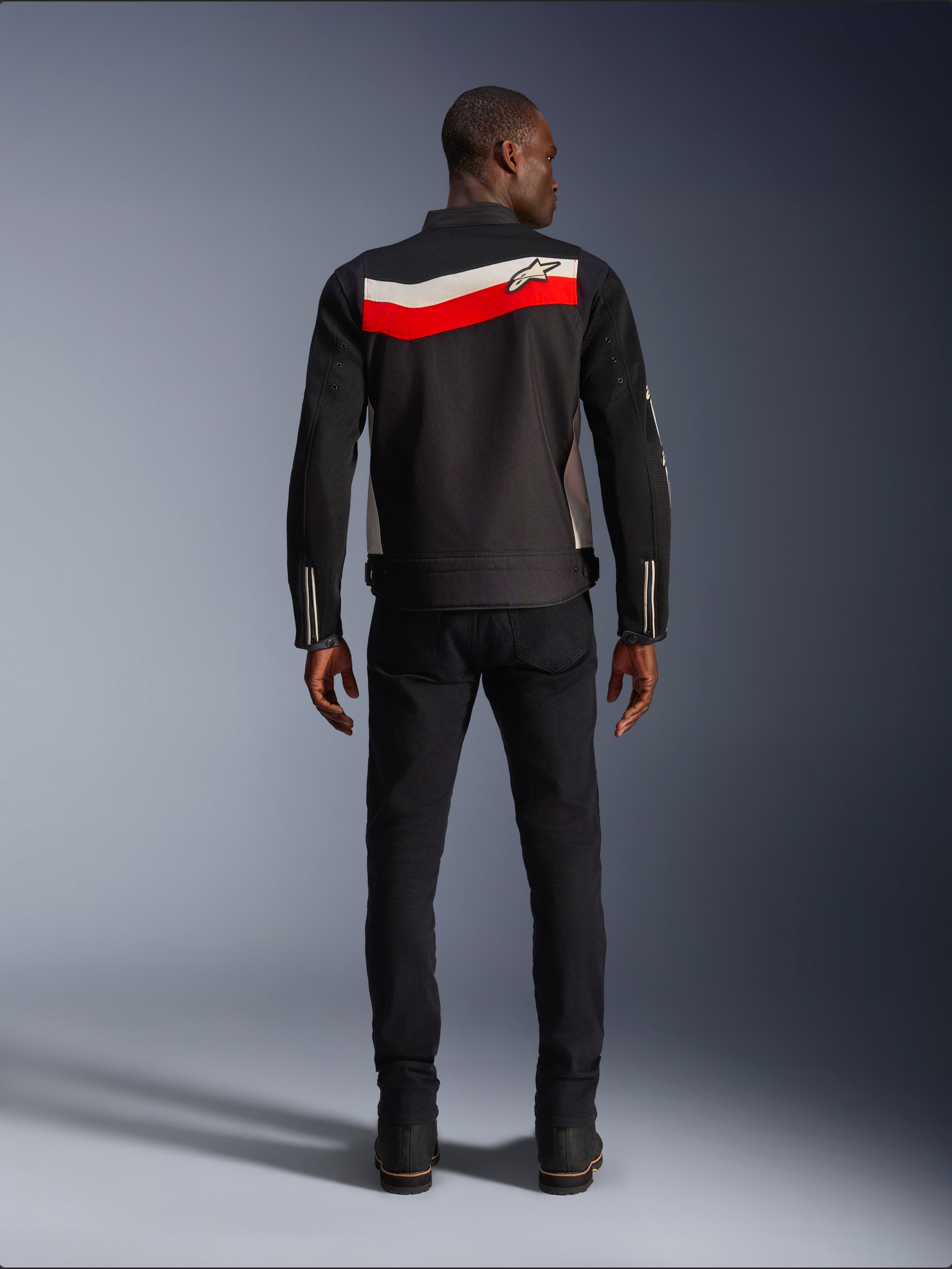 Veste Alpinestars T-Dyno Air, veste de moto textile pour homme, vue arrière, noire avec bandes horizontales rouge vif et écru dans le dos, logo Alpinestars sur l'empiècement, portée avec un pantalon et des bottes noirs, construction en maille 3D respirante