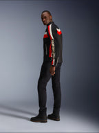 Un homme portant la veste Alpinestars T-Dyno Air en noir, rouge vif et écru, veste de moto en textile d'inspiration vintage avec des panneaux en maille, logo imposant sur la manche, associée à un pantalon et des bottes noirs pour un look motard moderne