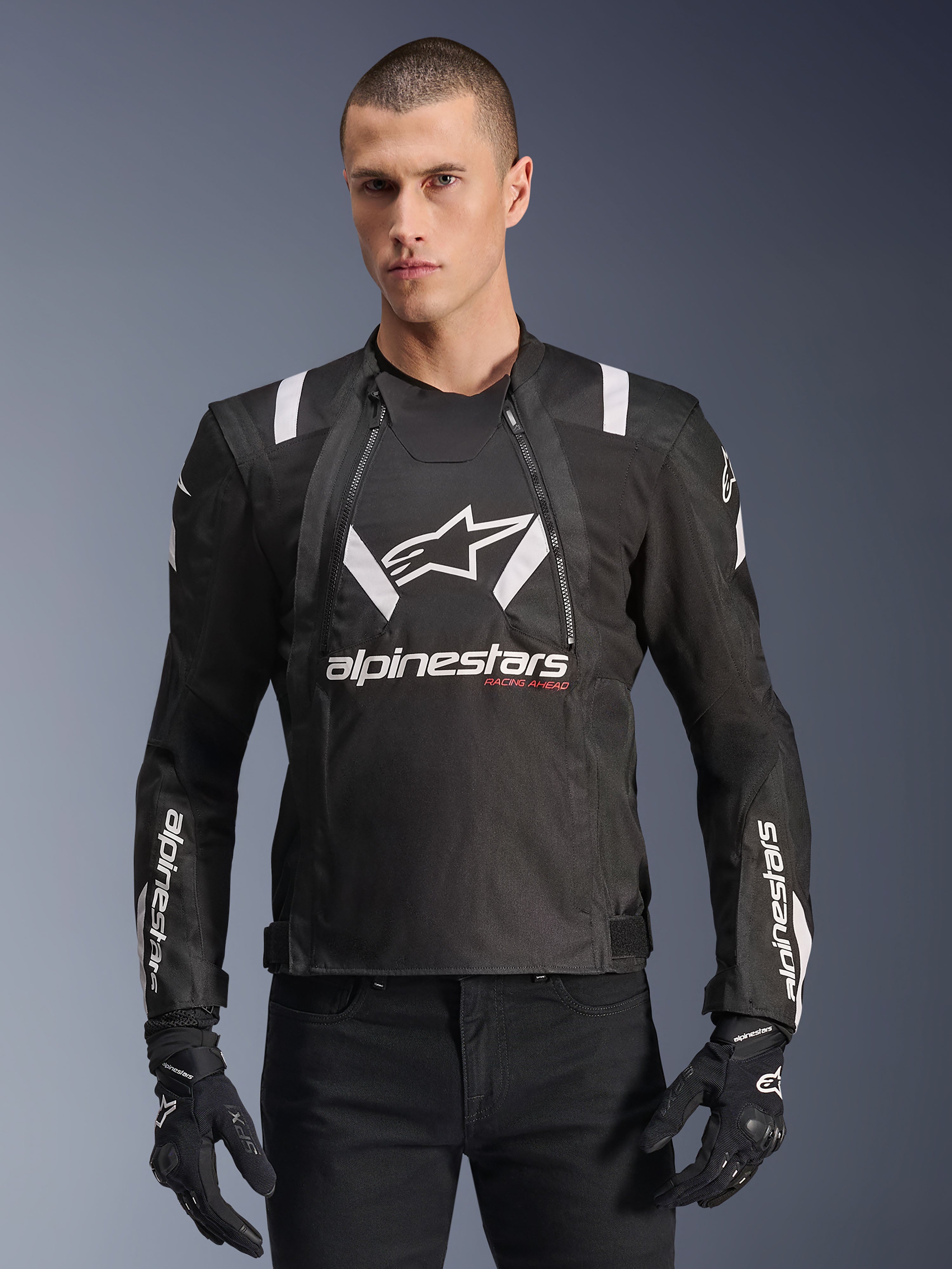 Alpinestars T-Stunt Air Jacket, veste de moto noire et blanche, portée par un homme avec des gants et un pantalon noirs, arborant des logos blancs sur la poitrine et les manches avec des accents rouges « Racing Ahead », design sportif épuré avec des bandes blanches sur les épaules et des panneaux en mesh respirant.