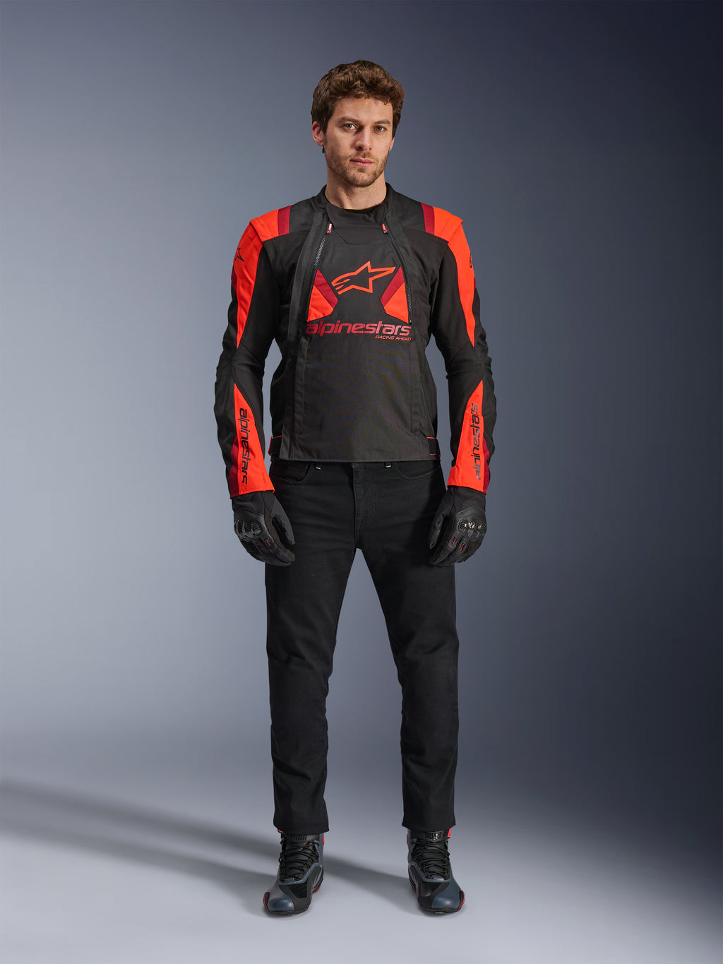Alpinestars T-Stunt Air Jas, zwart, fluo rood en donkerrood, gedragen door een model met een zwarte motorjeans en handschoenen, voorzien van een modulair 2-in-1 ontwerp met opvallende mesh-panelen voor luchtstroom, sportief gesneden motorkleding voor op de weg.