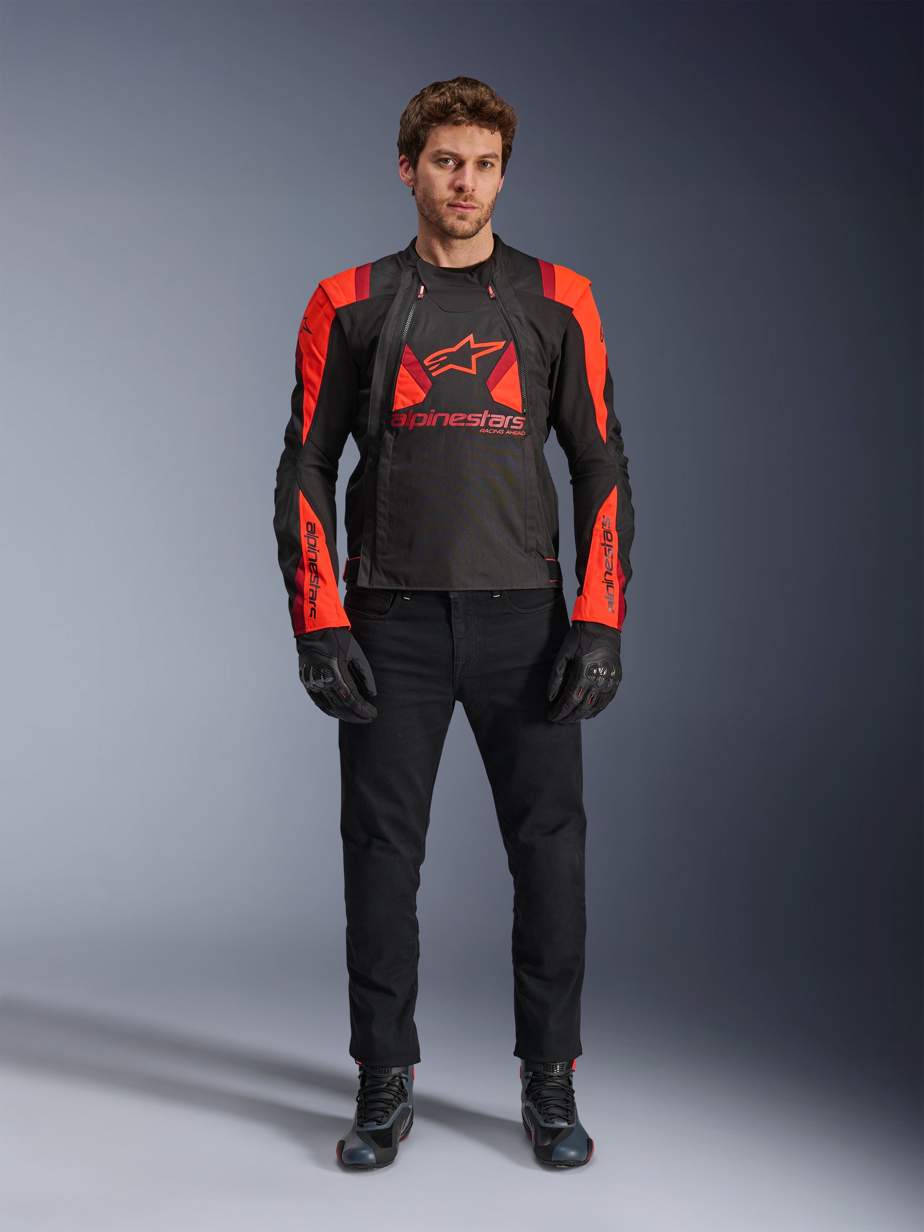 Alpinestars T-Stunt Air Jas, zwart, fluo rood en donkerrood, gedragen door een model met een zwarte motorjeans en handschoenen, voorzien van een modulair 2-in-1 ontwerp met opvallende mesh-panelen voor luchtstroom, sportief gesneden motorkleding voor op de weg.